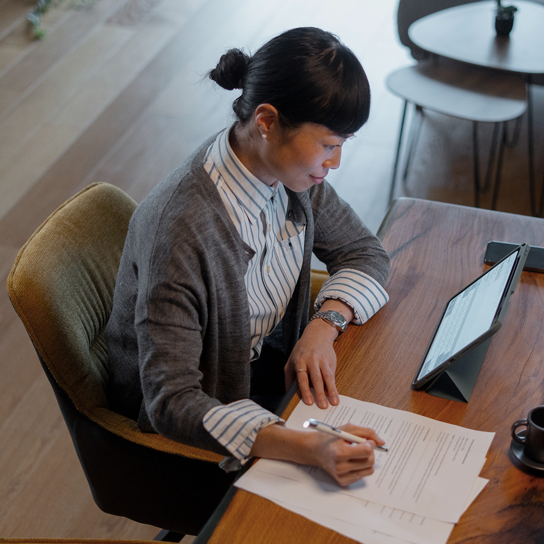 Woman writes on paper documents at a wooden desk with a tablet and coffee cup nearby