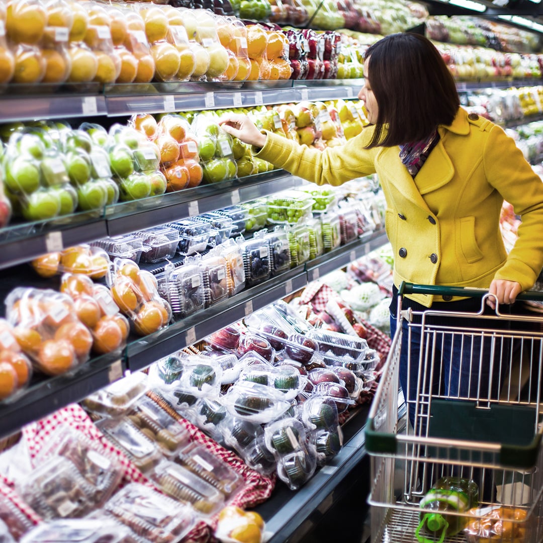 A woman in a yellow coat pushes a shopping cart while selecting green apples from a well-stocked produce aisle in a modern supermarket.