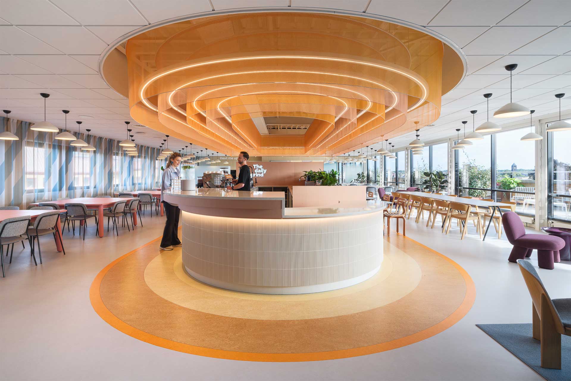 Een modern, ruim café met een centrale, ronde toonbank onder gelaagde, oranje lichtgevende plafondpanelen. Twee mensen staan bij de toonbank te praten. Grote ramen laten natuurlijk licht binnen, waardoor een uitnodigende sfeer ontstaat.
