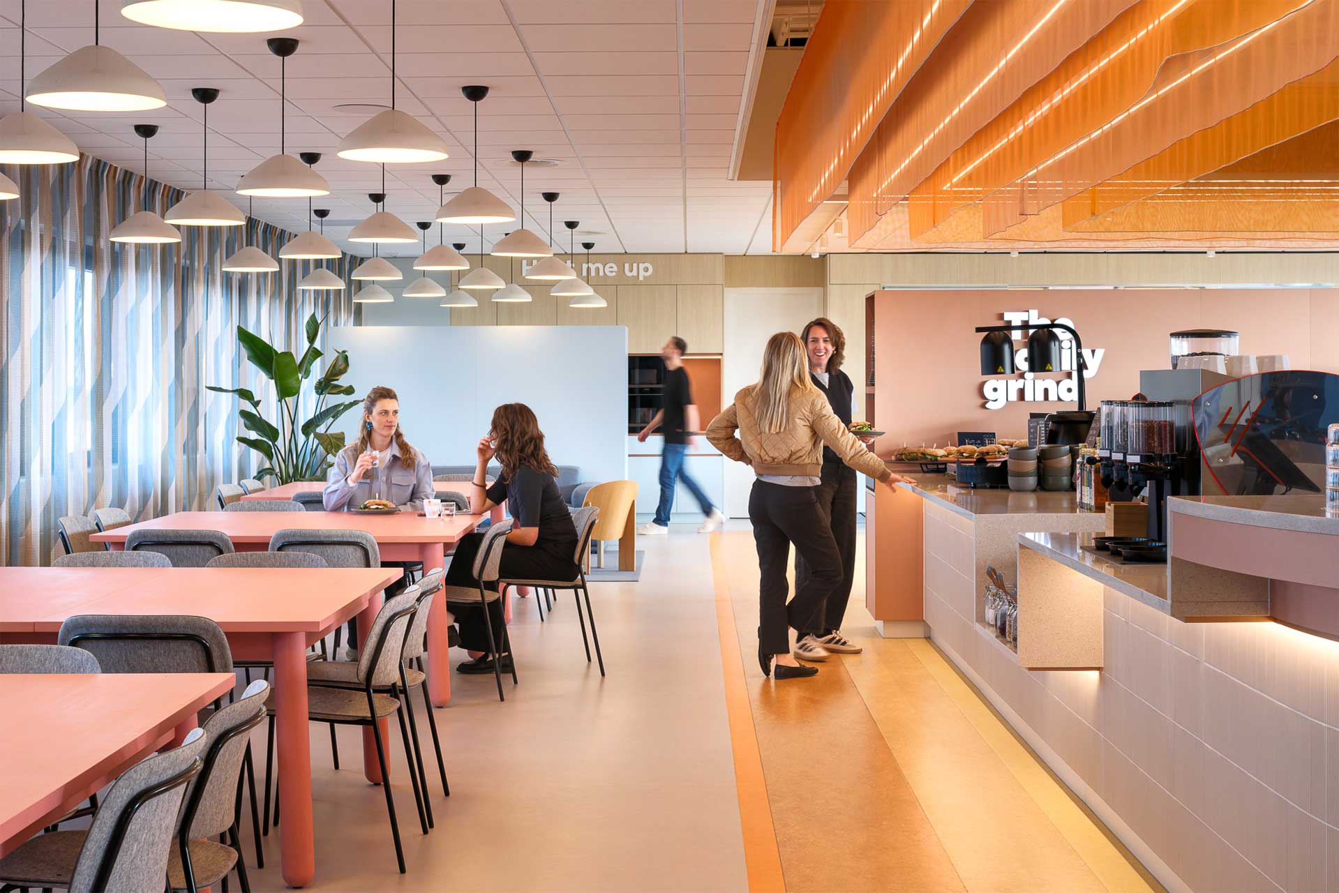 Moderne cafetaria met roze tafels, zwarte stoelen en hanglampen. Mensen zijn aan het socializen en eten aan het uitkiezen. De sfeer is gastvrij en levendig.
