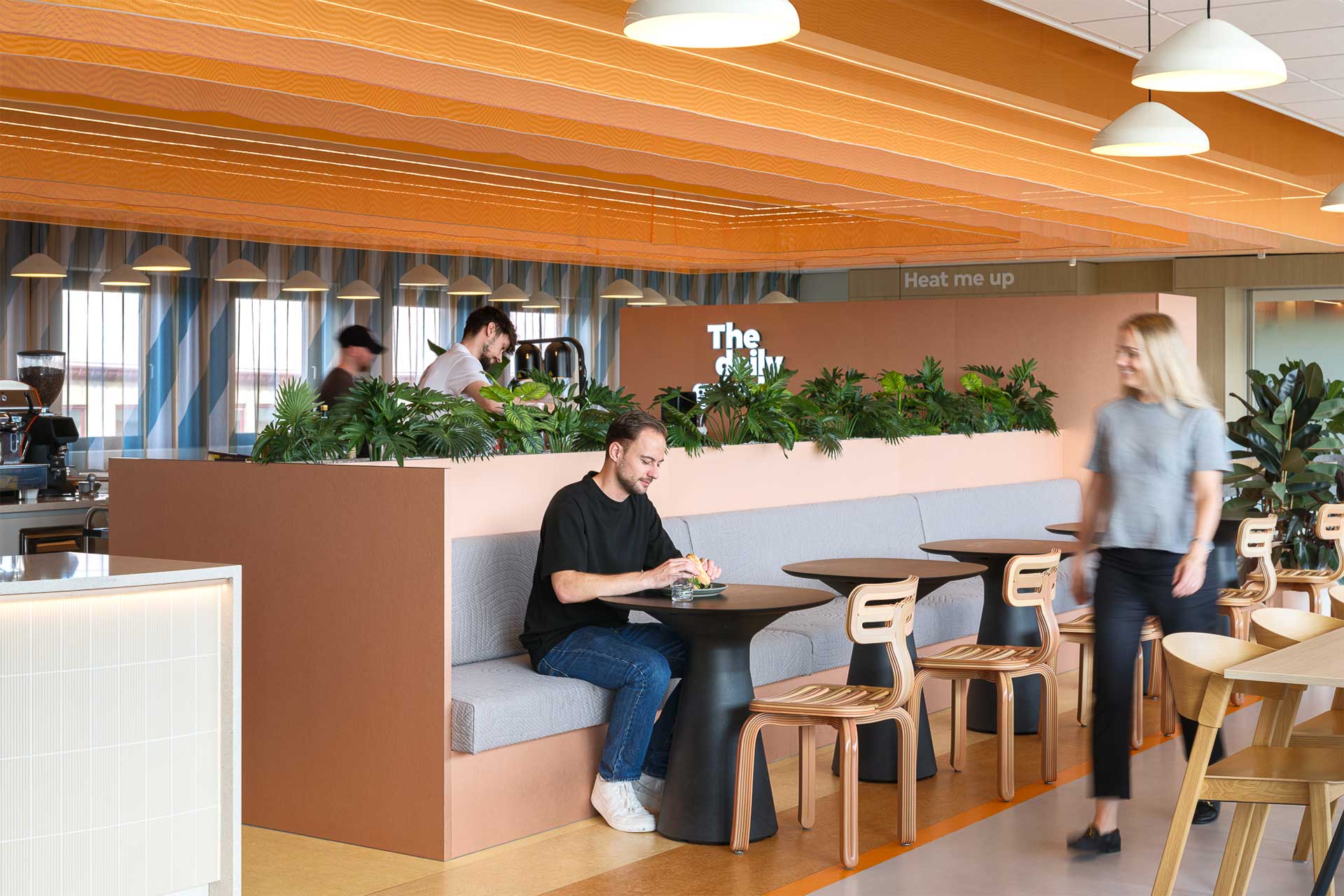 Een modern café met zachte verlichting en houten accenten toont een man die alleen zit te eten in een hoekje met veel groen. Mensen lopen rond, wat er voor zorgt dat er een ontspannen sfeer hangt.