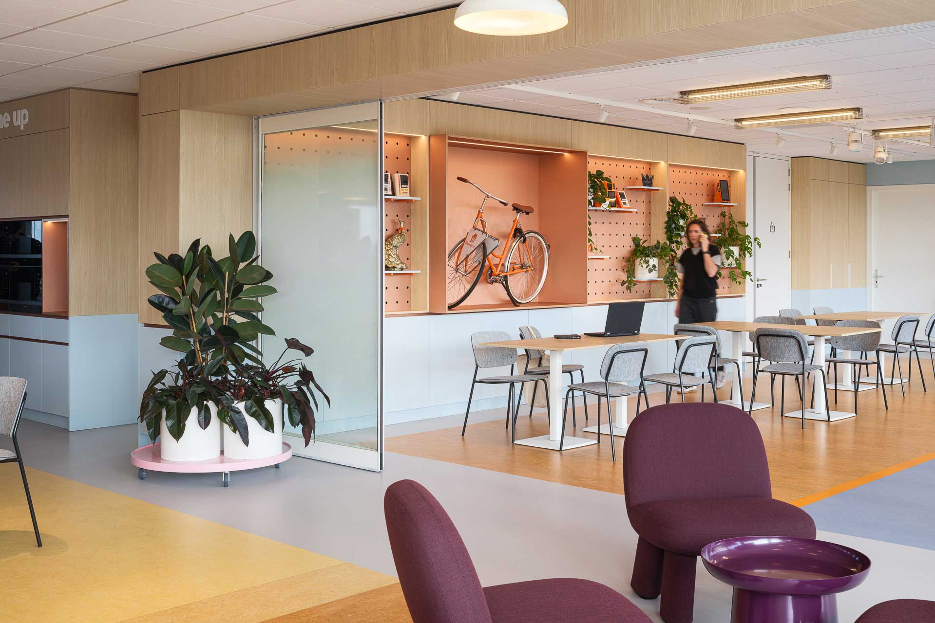 Moderne kantoorlounge met een houten tafel, grijze stoelen en een aan de muur bevestigde oranje fiets. Groene planten zorgen voor frisheid. Een persoon staat bij de planken.
