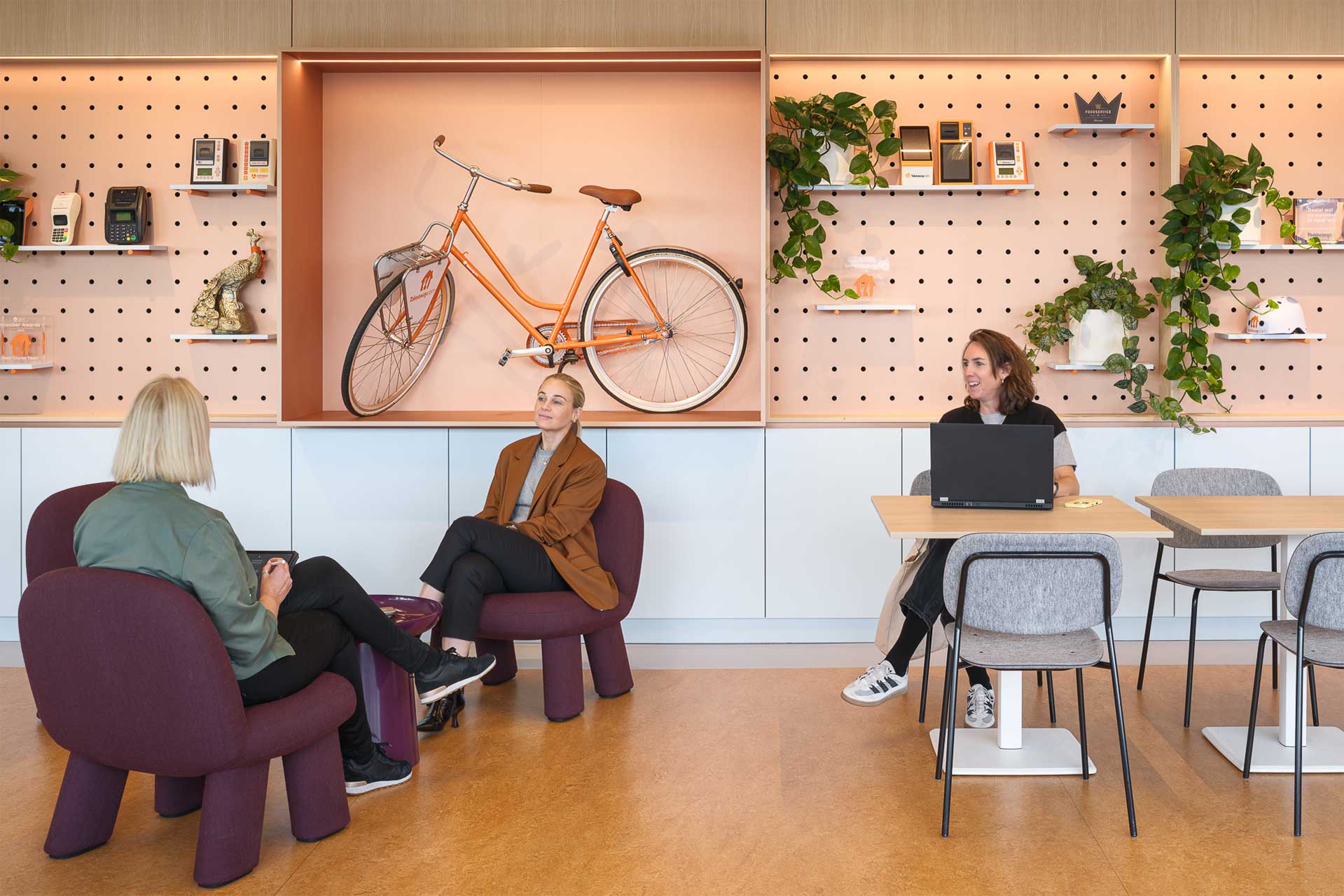 Drie mensen in een moderne kantoorlounge met houten planken en een oranje fiets in de vitrine. Groene planten en een stijlvolle inrichting zorgen voor een ontspannen, creatieve sfeer.