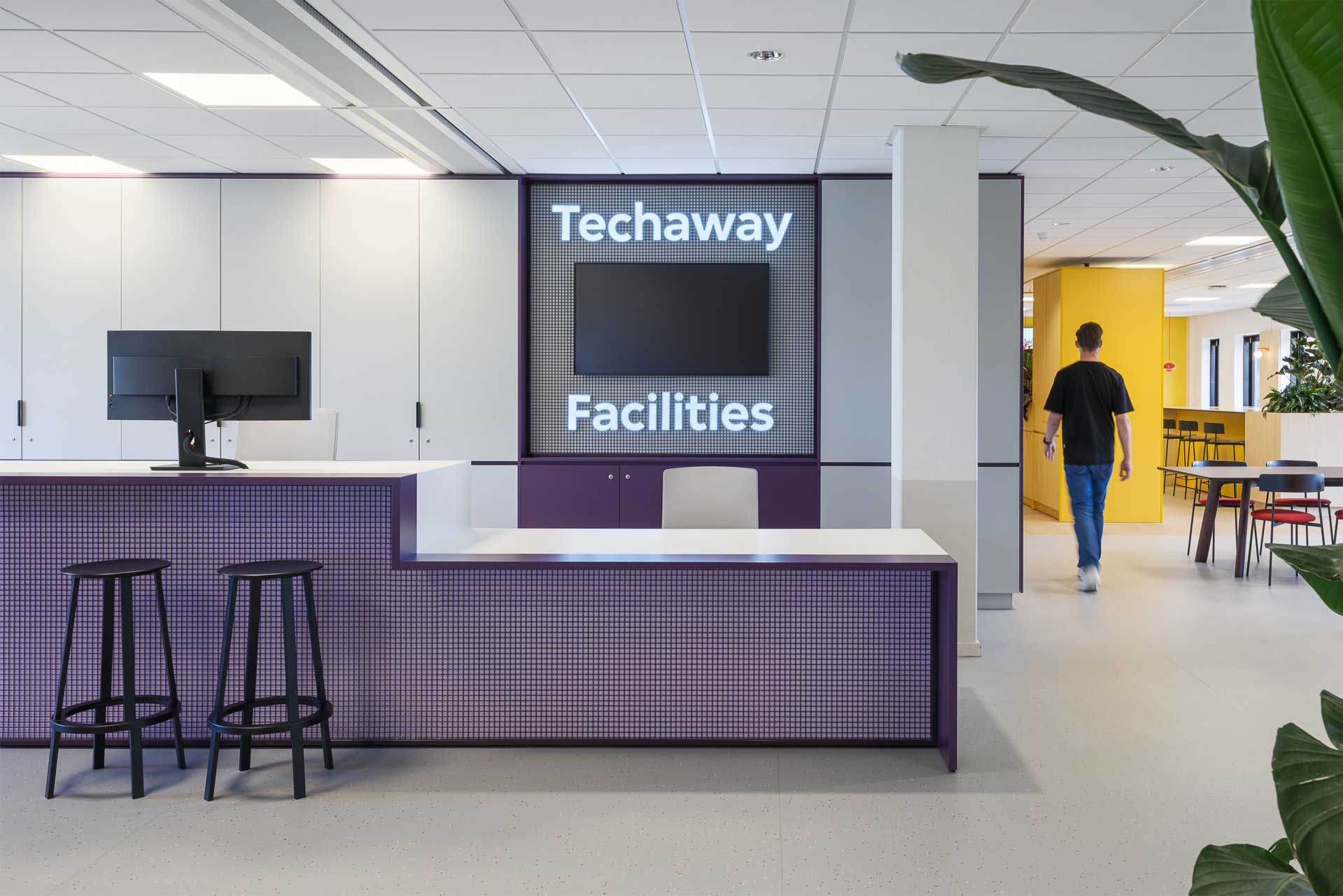 Moderne kantoorlobby met een paarse receptiebalie, twee zwarte krukken, een monitor en ‘Techaway Facilities’ aan de muur. Een persoon loopt naar een gele gang.