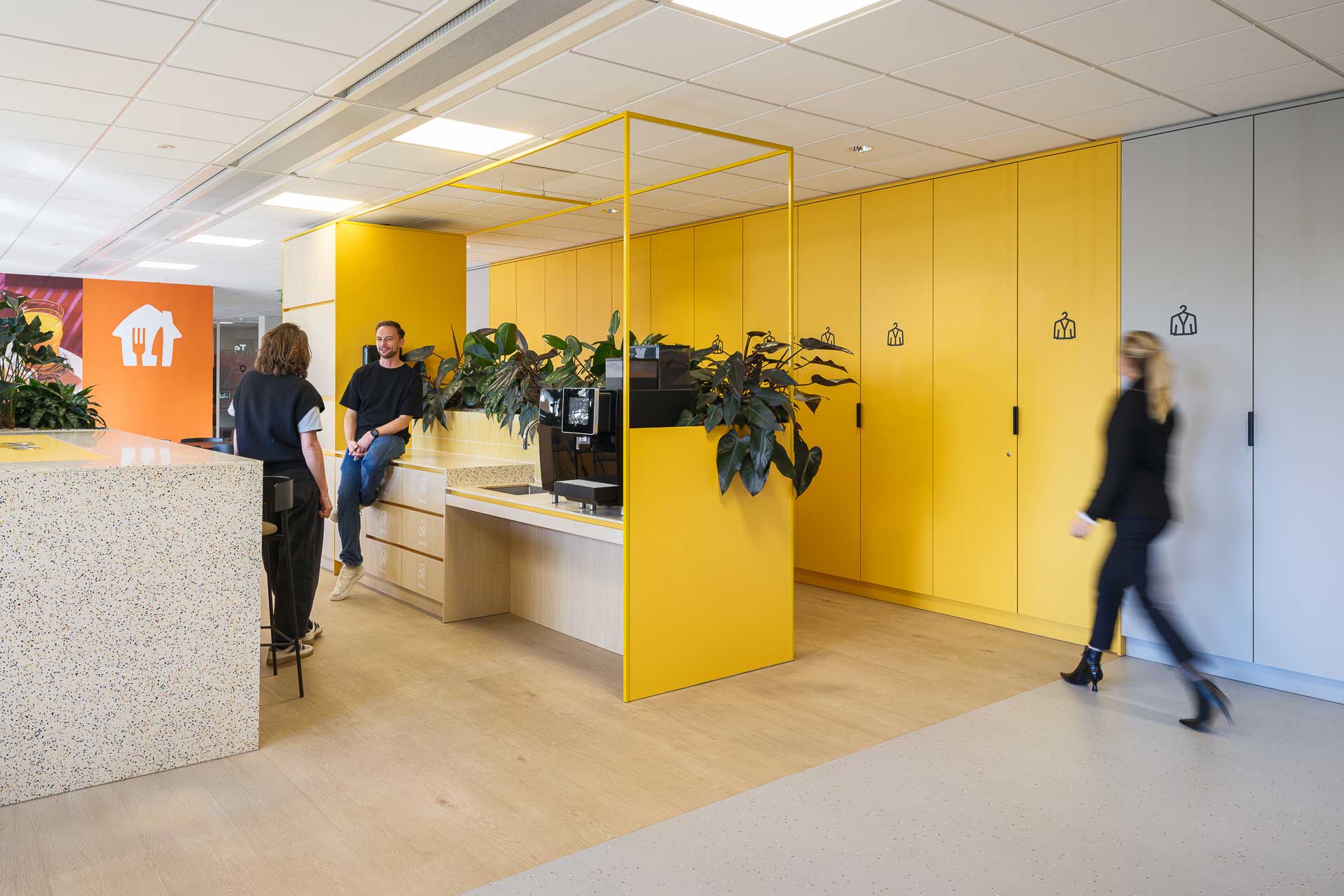 Moderne kantoorruimte met gele kasten, planten en terrazzo oppervlakken. Twee mensen praten ontspannen met elkaar, terwijl een ander voorbij loopt, wat een levendige sfeer zorgt.