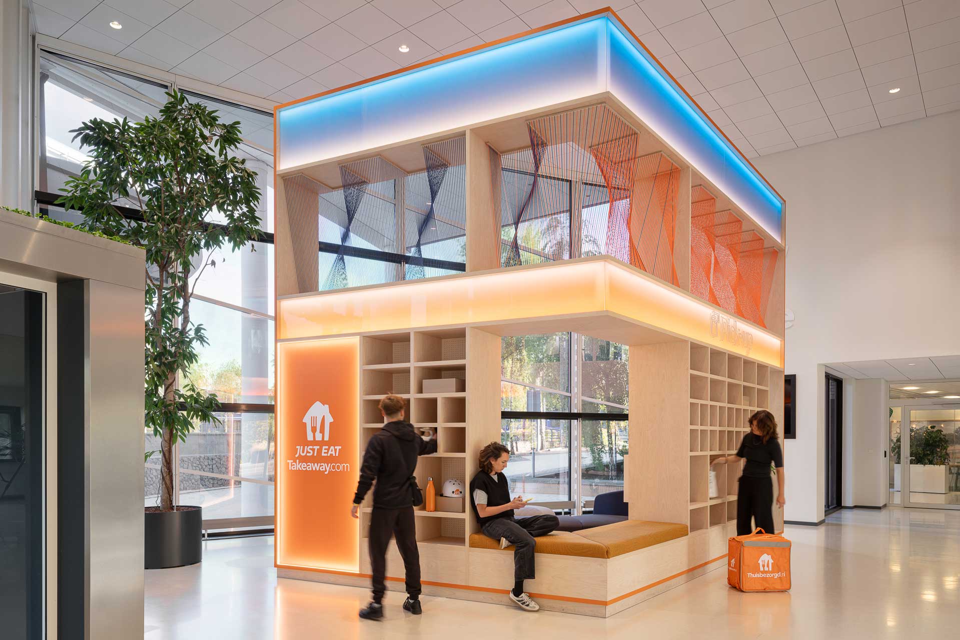 Moderne lobby met een lounge van twee verdiepingen hoog met een verlicht ‘Just Eat Takeaway’-logo. Drie mensen praten met elkaar in een lichte, ruime ruimte met grote ramen.