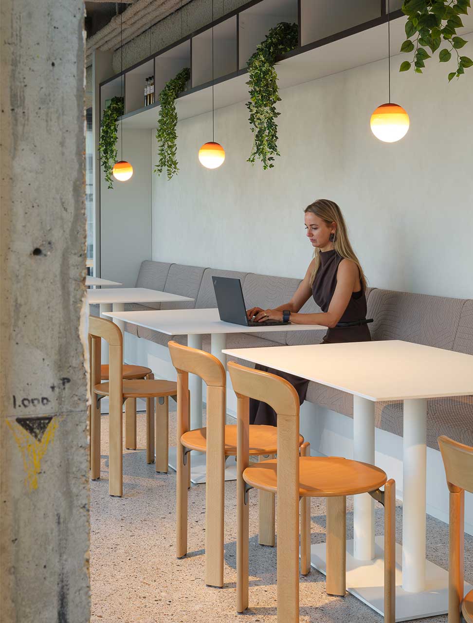 A woman works on a laptop at a modern cafe with white tables, wooden chairs, and hanging plants. Warm pendant lights create a cozy atmosphere.