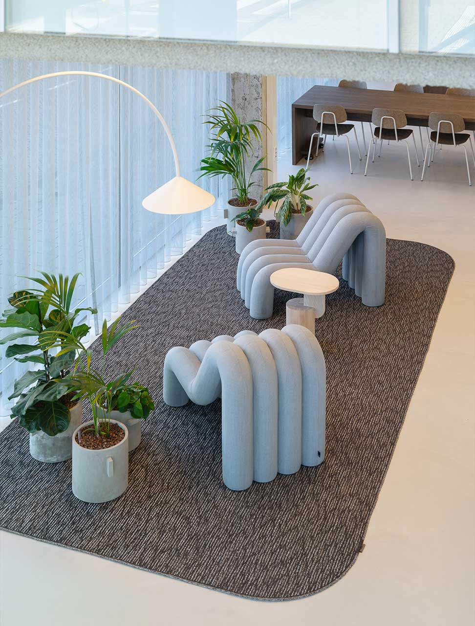Modern interior with curved gray chairs on a dark textured rug, large potted plants, and minimalist white lamp. A dining area is visible in the background.