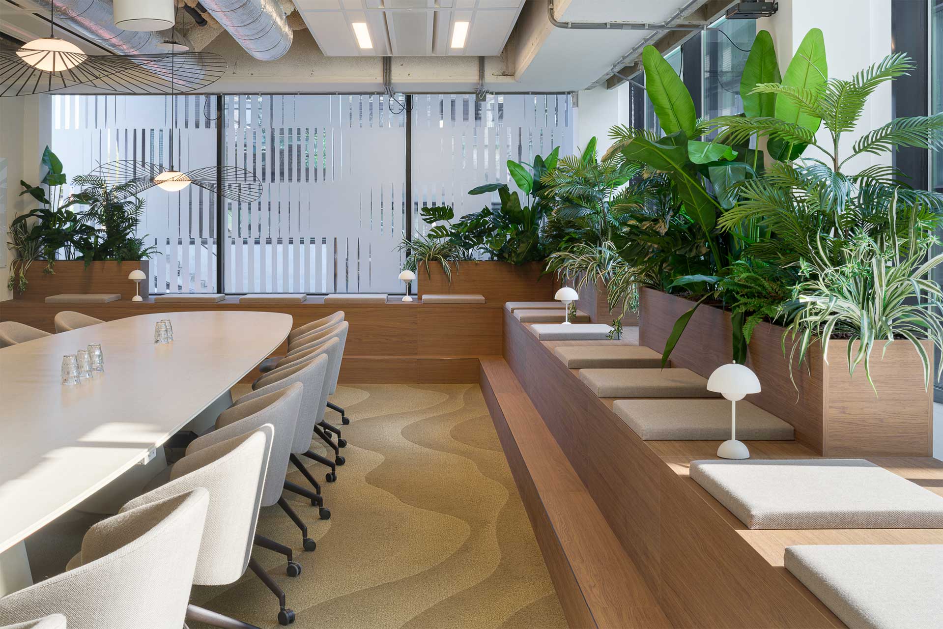 Modern meeting room with a long table and cushioned chairs beside tiered seating. Lush green plants and ambient lighting create a fresh, calming atmosphere.