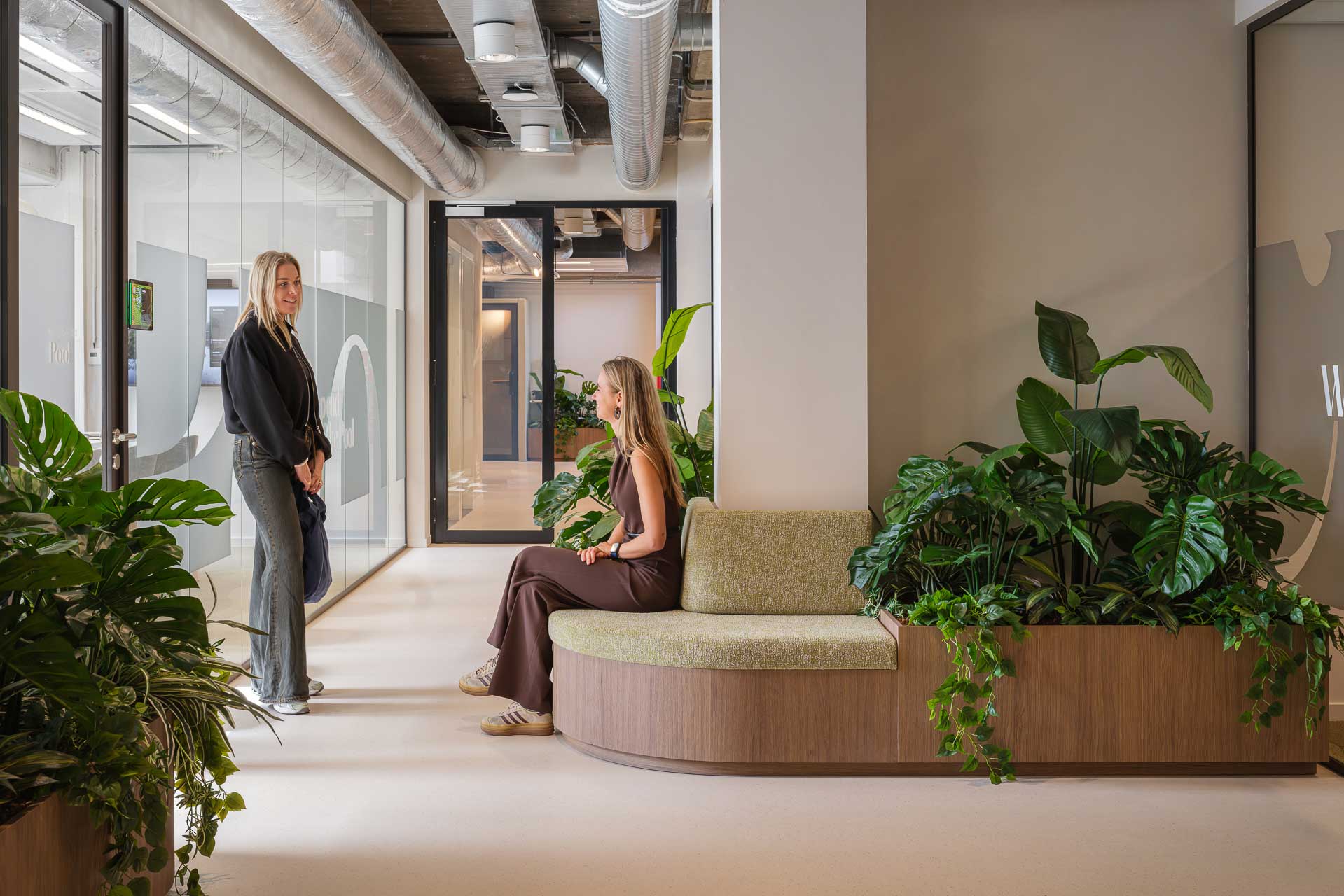 Two women in a modern office lobby with green plants. One sits on a cushioned bench, while the other stands nearby. The setting is calm and contemporary.