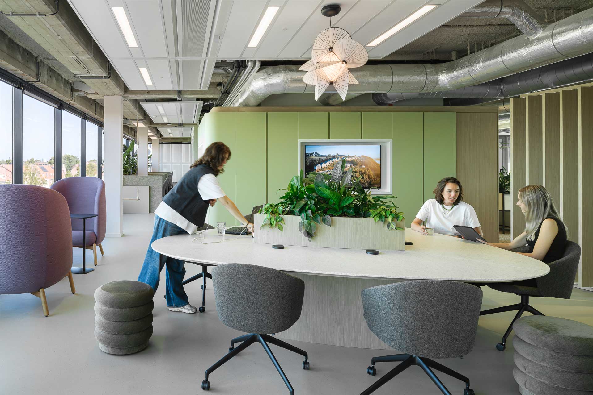 Modern office space with three people around an oval table, featuring a central plant arrangement. Large windows bring in natural light, creating an airy, collaborative atmosphere.