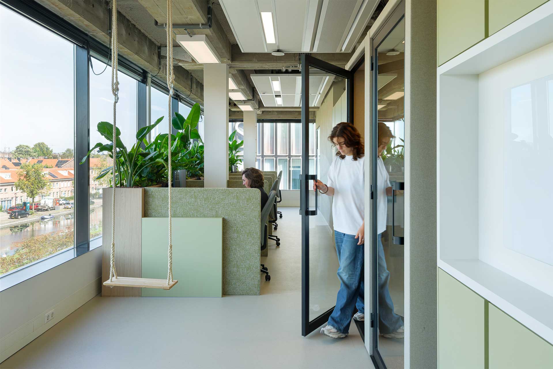 An office with large windows and lots of greenery. One person opens a glass door, while another sits at a desk.