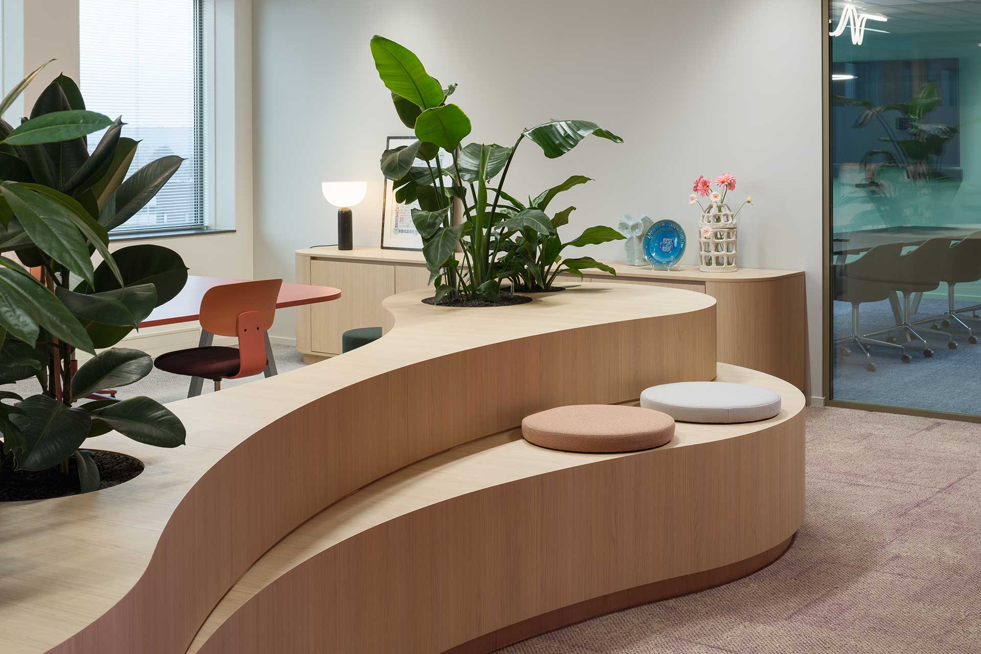 Modern office space with undulating wooden seating element, green plants, soft seat cushions and adjoining meeting room.