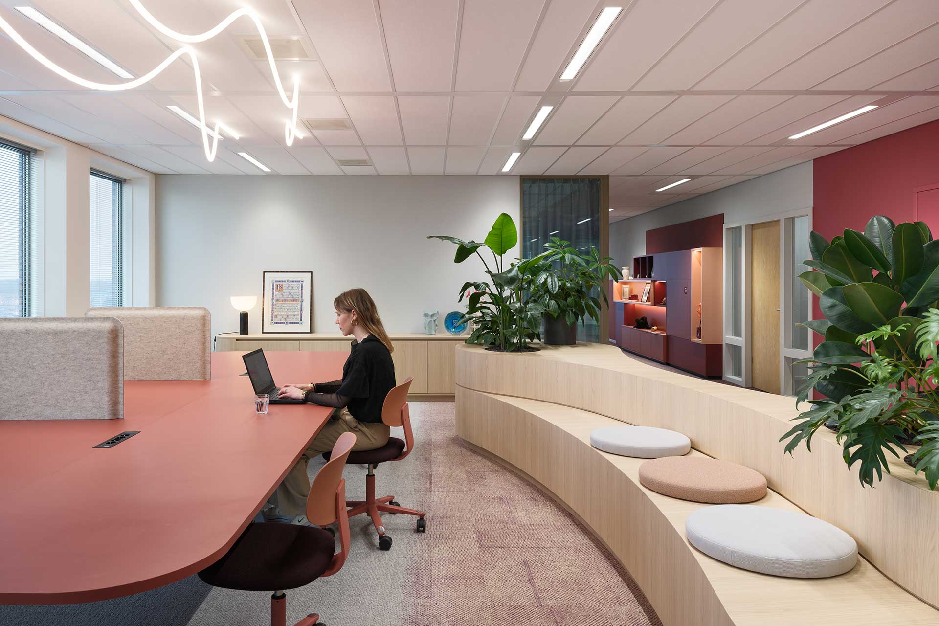 Modern office space with shared work table, felt privacy screens, wooden seating element with seat cushions and lots of green plants.