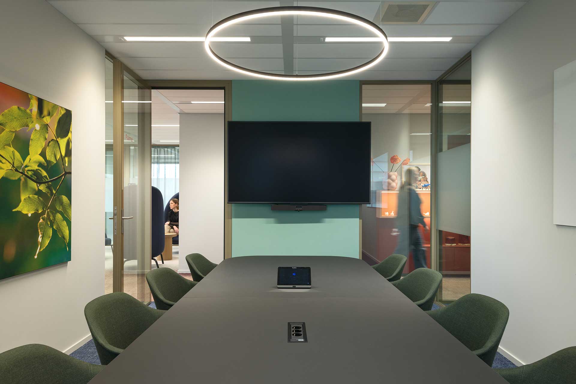 Modern meeting room with long conference table, large monitor, round designer ceiling lamp and glass walls to adjacent offices.