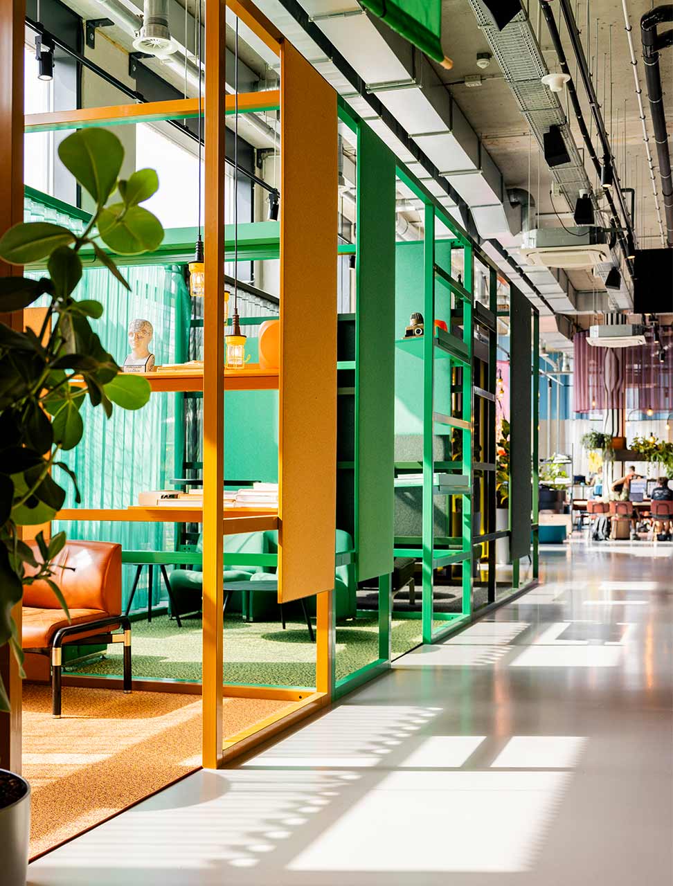 Open-plan office with industrial design, colorful acoustic partitions, indoor plants, modern seating areas, and natural daylight from skylights or windows.