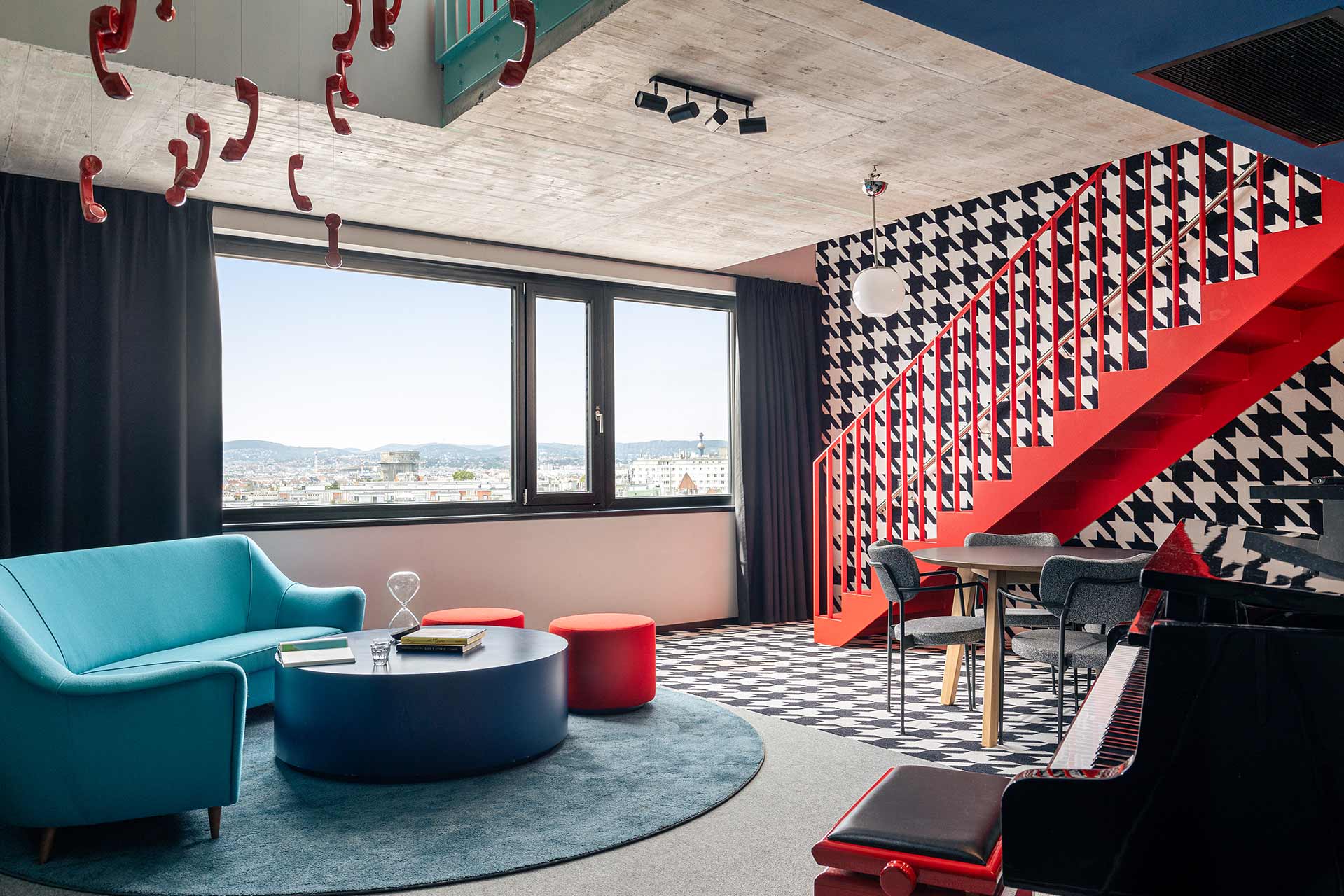 Modern living room with blue sofa, round coffee table, red pouffes, striking red staircase and large window overlooking the city.
