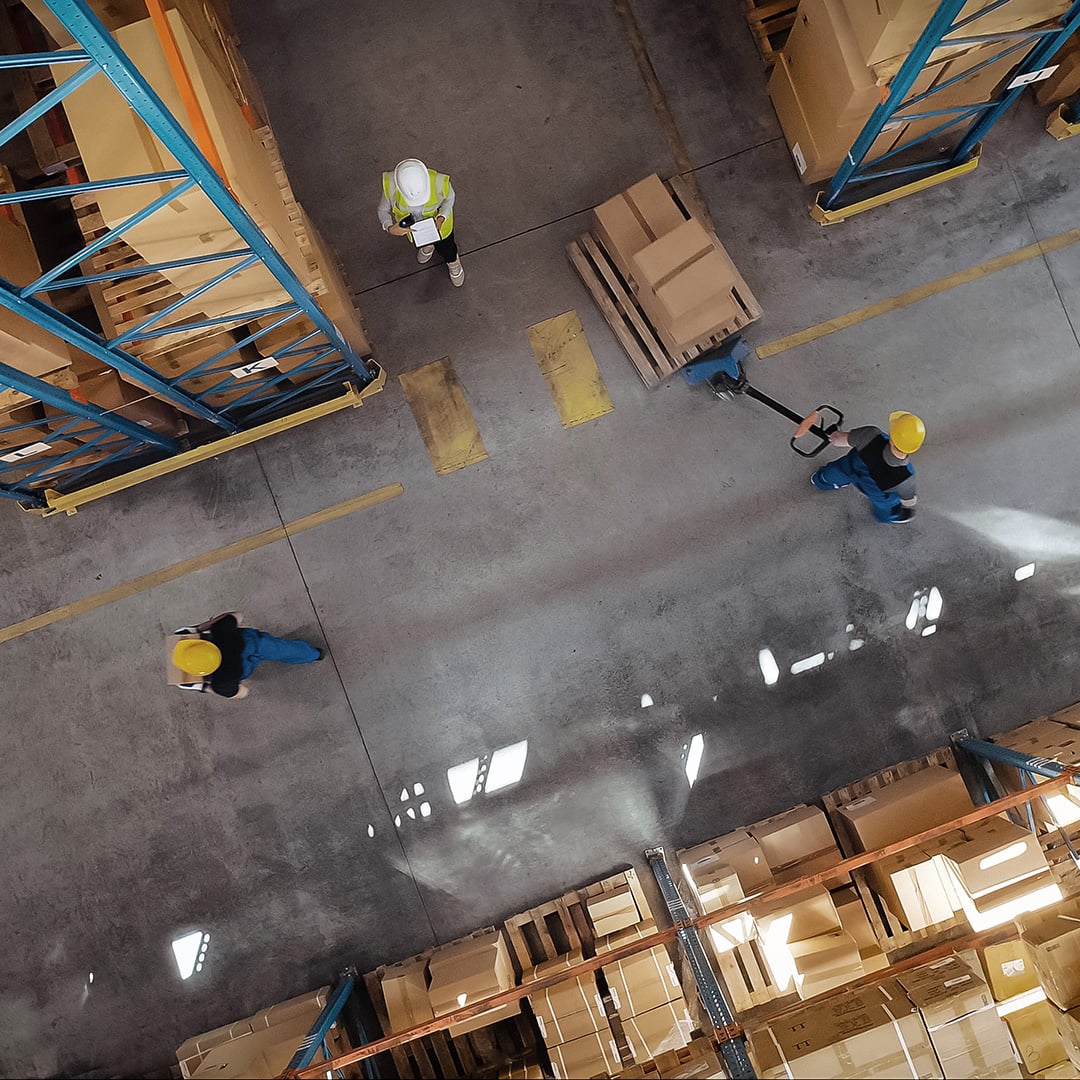 Overhead view of a warehouse aisle with workers moving pallets and boxes between tall storage racks.