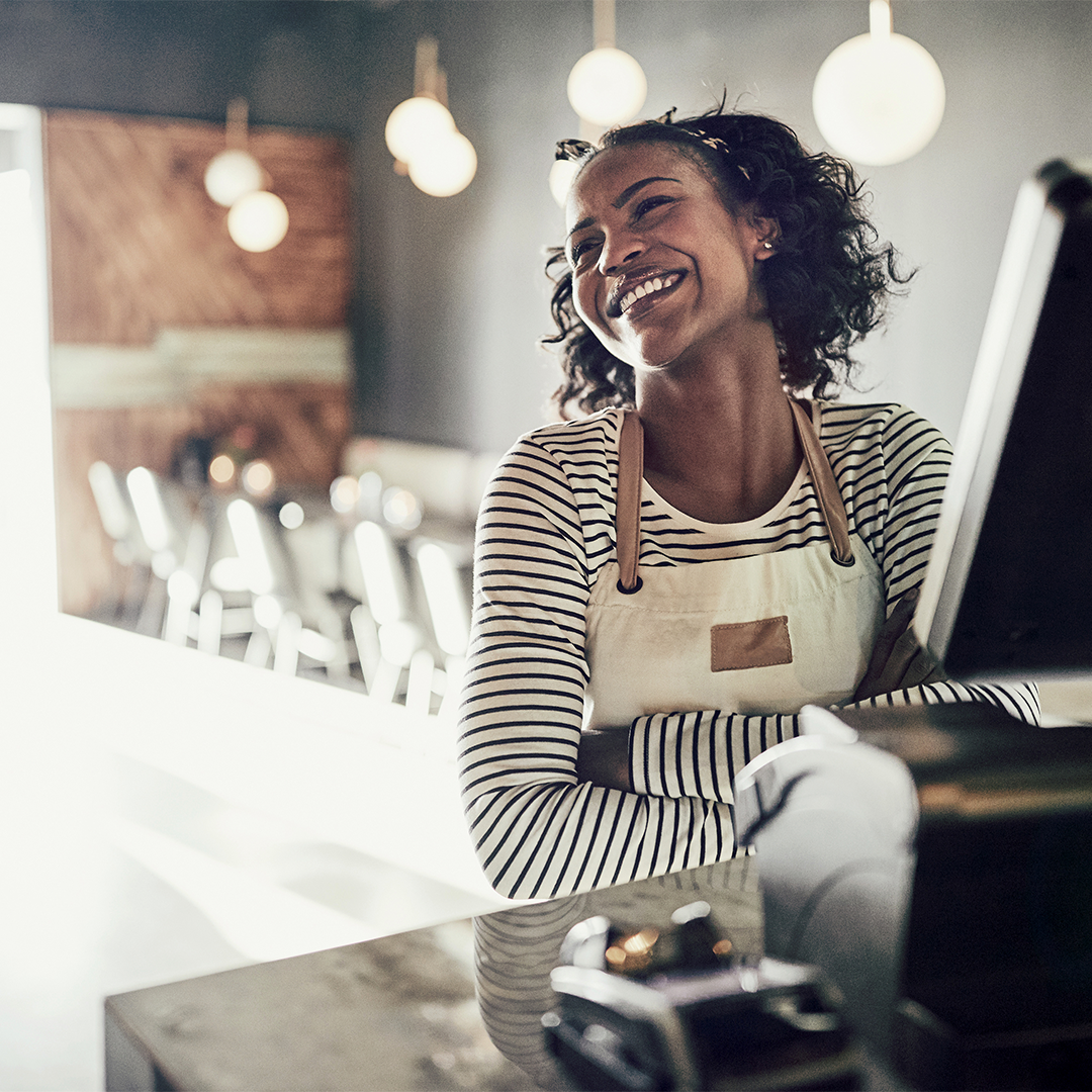 A smiling woman in an apron stands behind a counter in a modern, well-lit cafe, representing a successful small business in a commercial space.