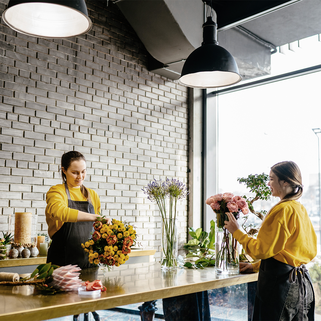 Two women arranging flowers in a modern flower shop with a brick wall and large windows.