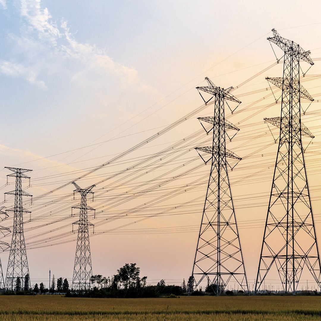 High‑voltage transmission towers stretch across open fields beneath a colorful sky at sunset.