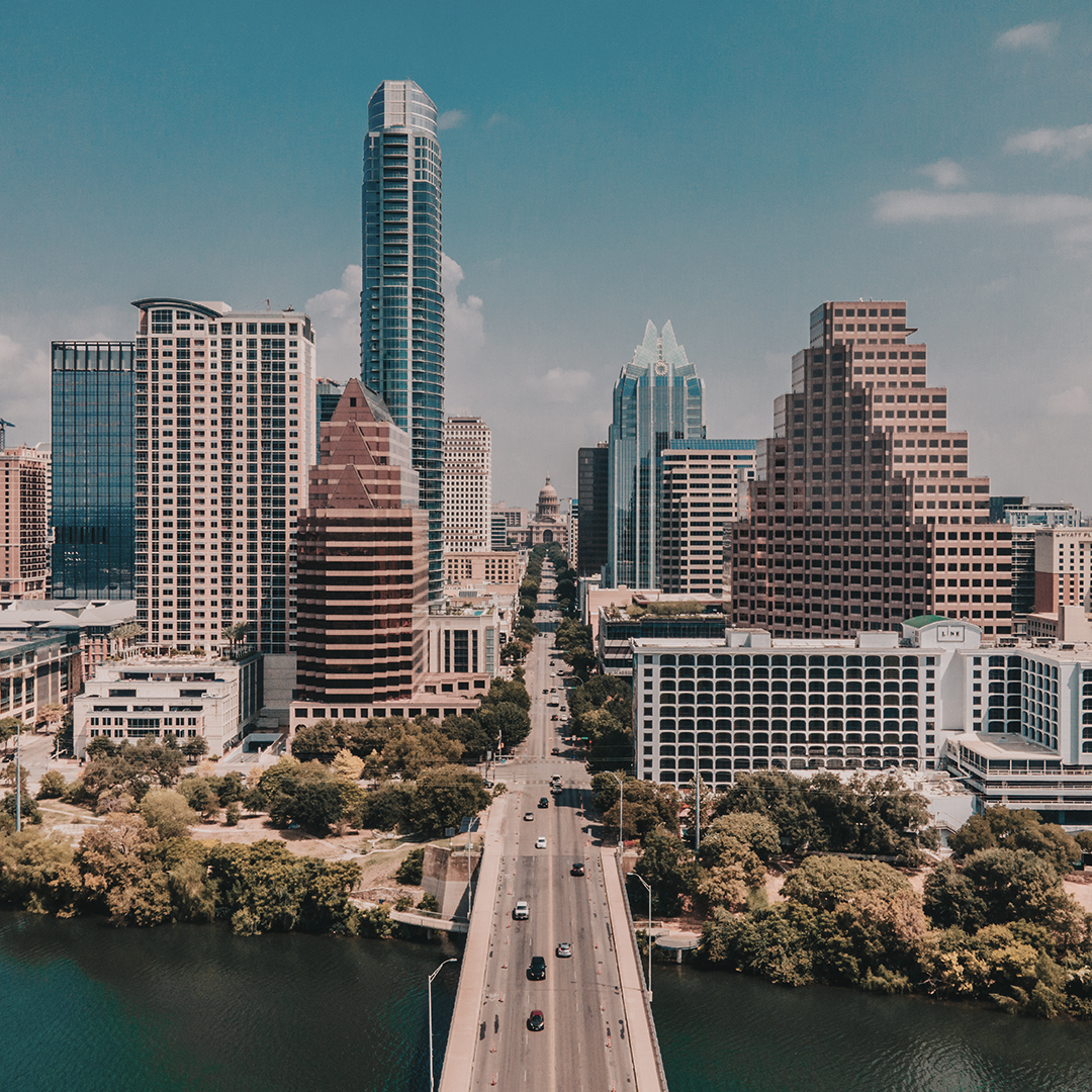 Cityscape with a central road crossing water into a skyline of modern glass and traditional stepped buildings under a partly cloudy sky.