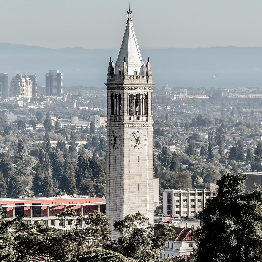 The Campanile (Sather Tower) at UC Berkeley with clock faces and a cityscape of buildings and trees in the background.
