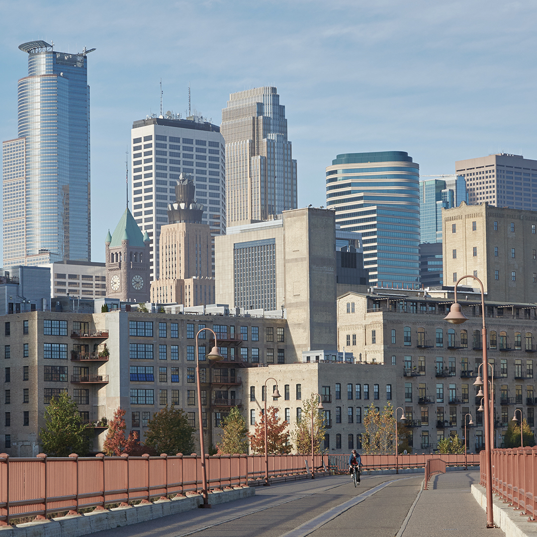 Cityscape with a cyclist crossing a red-railed bridge, surrounded by modern skyscrapers, older buildings, and autumn-colored trees.