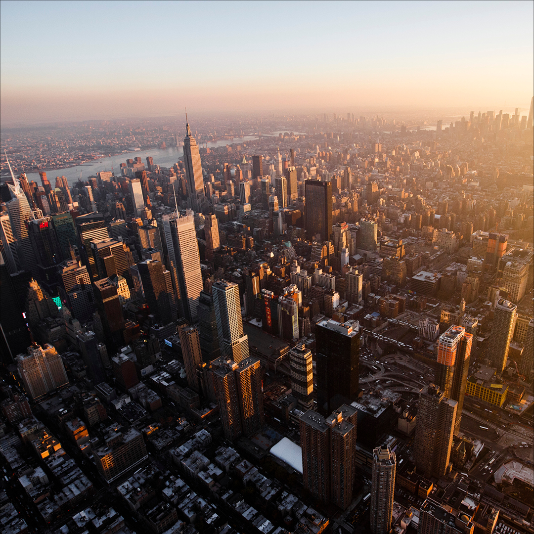 Aerial view of New York City at sunset featuring the Empire State Building, bridges over the river, and a golden glow across the skyline.
