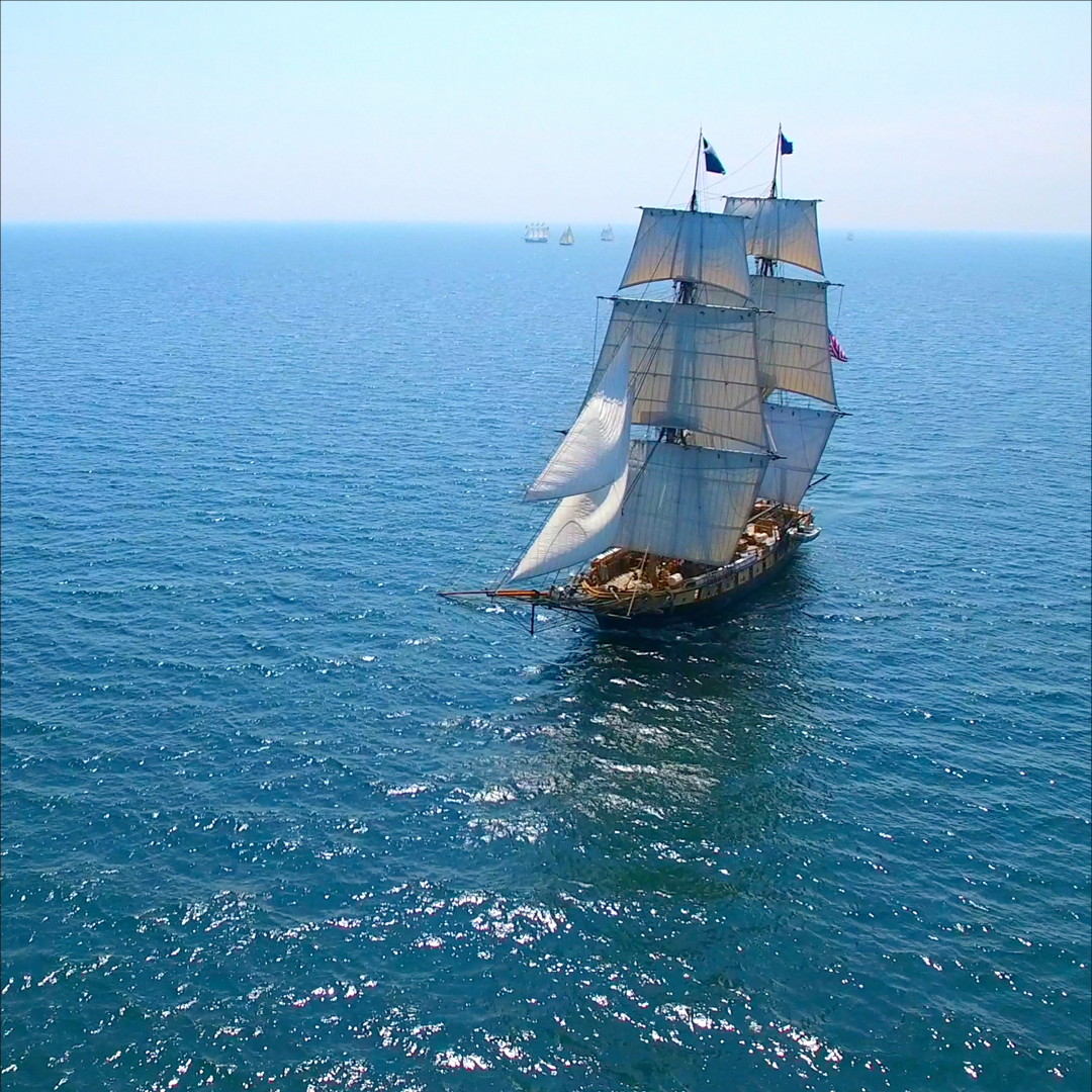 Historic tall sailing ship with full sails deployed, moving through calm blue sea with other ships on the horizon.