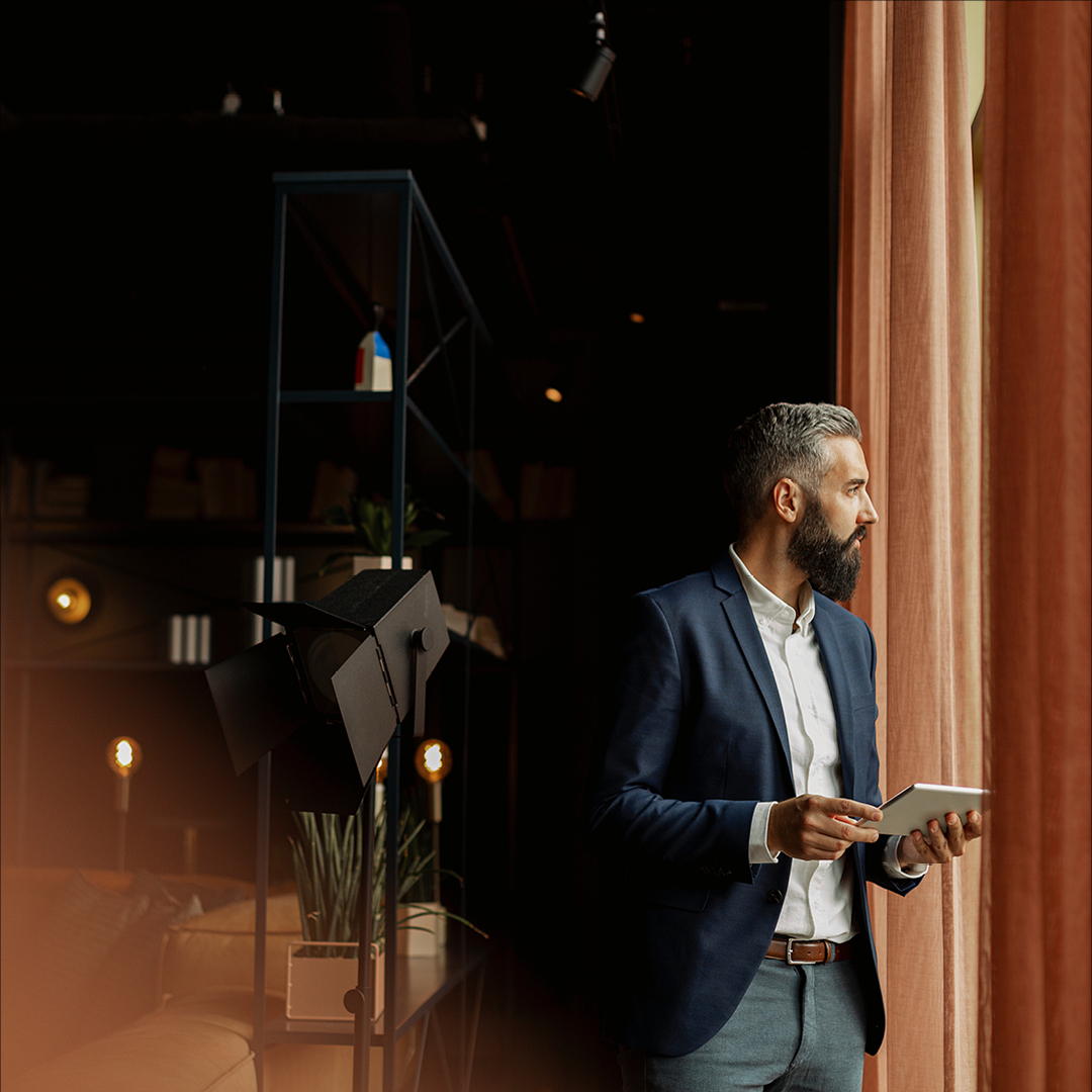 Person in a navy blazer holding a tablet near floor-to-ceiling curtains in a modern, warmly lit interior.