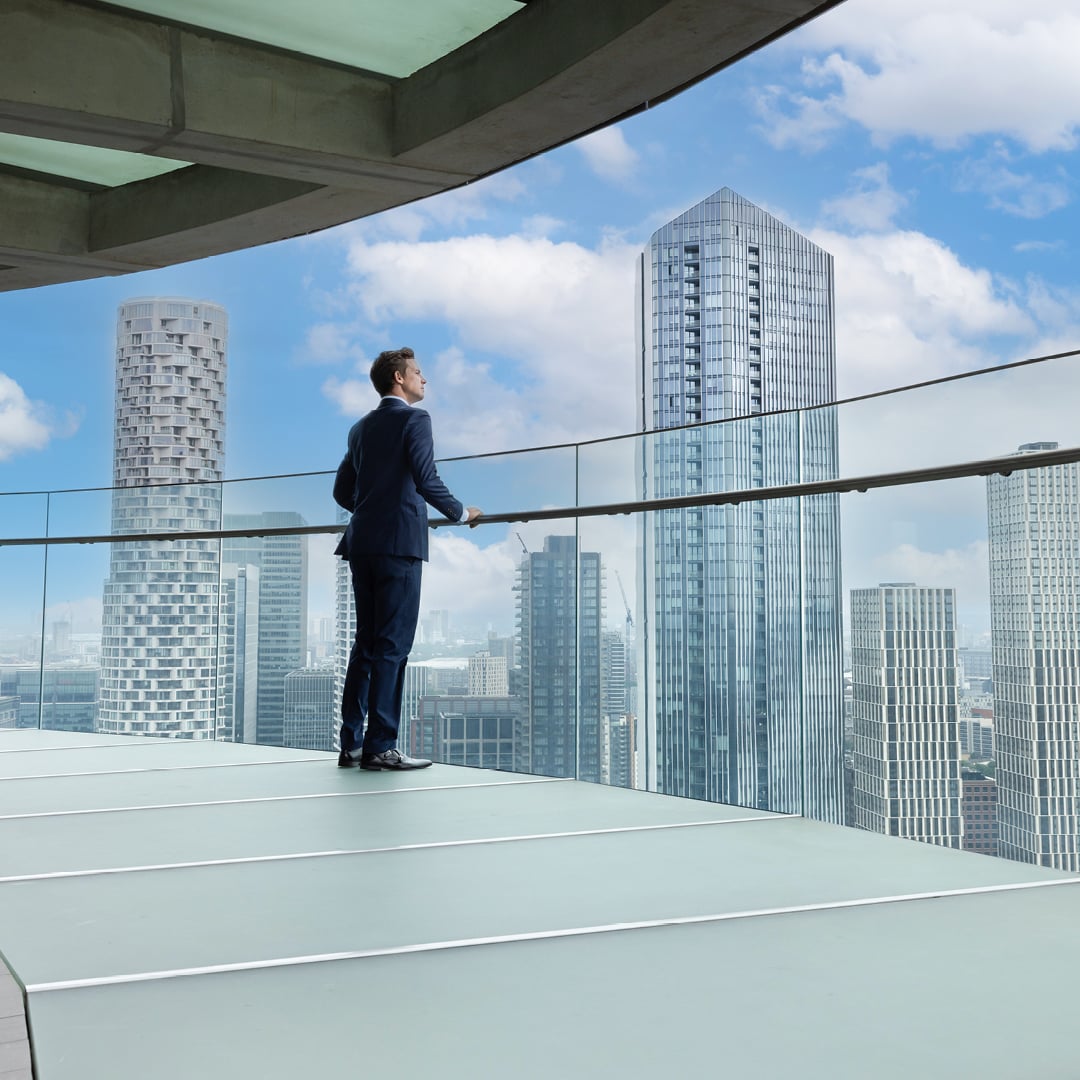 Person in a formal suit standing on a high-rise balcony overlooking a modern city skyline with glass skyscrapers.
