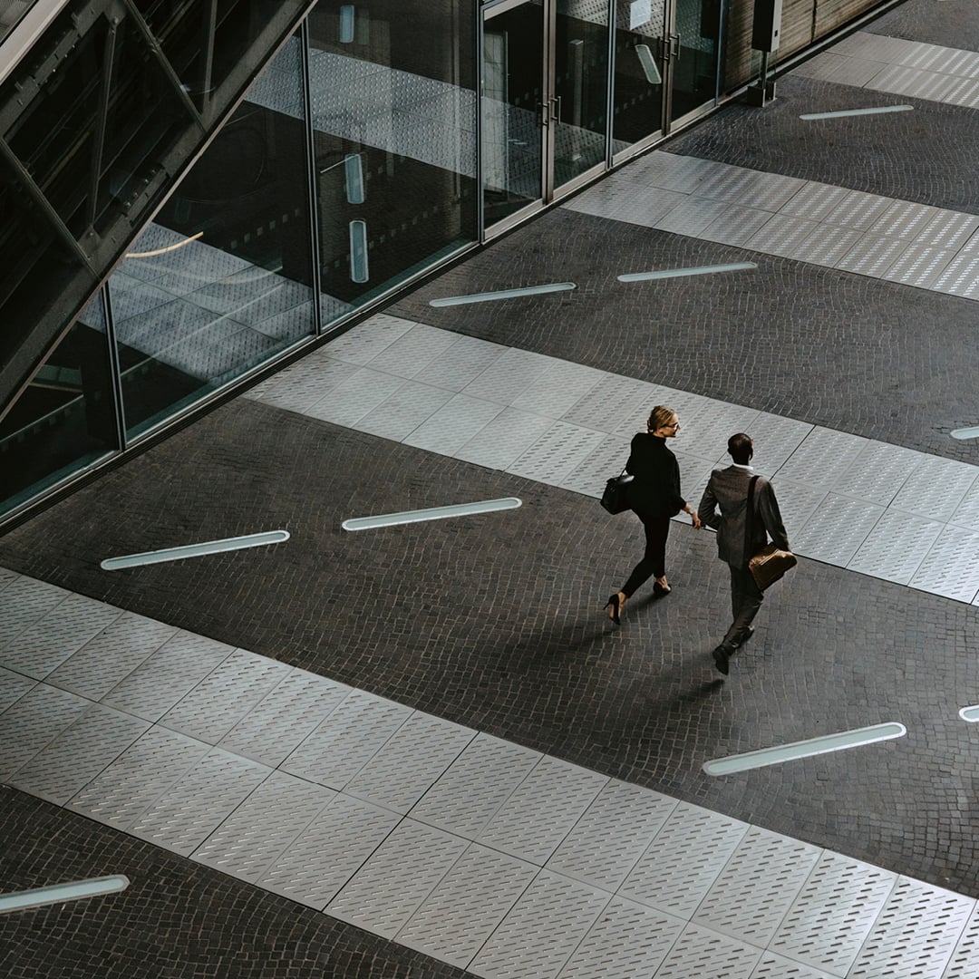 Two people walking across a patterned urban plaza beside a glass‑fronted building