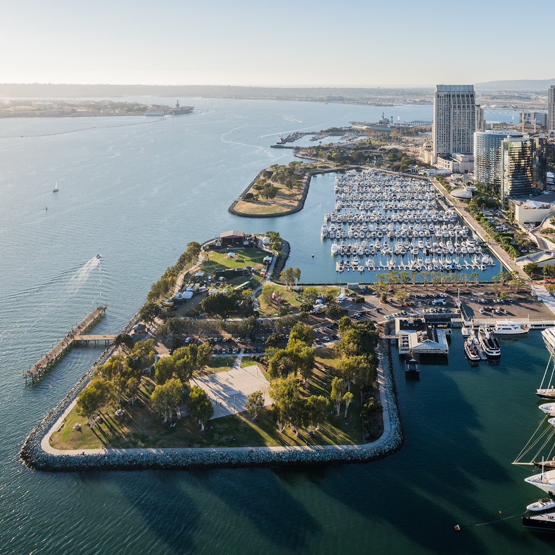 Aerial view of a waterfront district with a marina full of boats, surrounding parkland, walking paths and nearby office towers along a harbor under clear daylight.