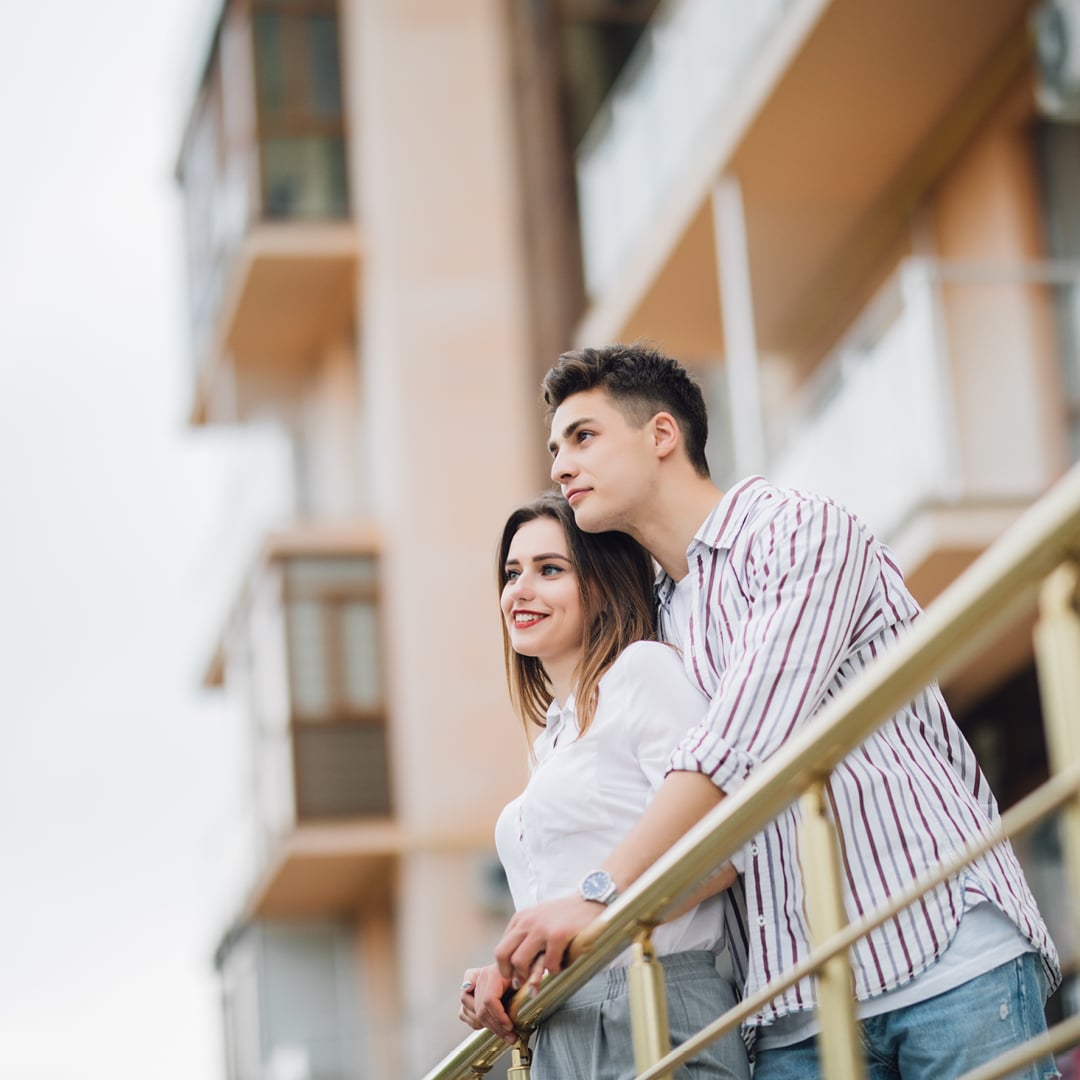 Young couple looking out from the balcony of a multifamily apartment building, illustrating workforce housing renter demand discussed by TruAmerica CEO Bob Hart on CBRE's The Weekly Take