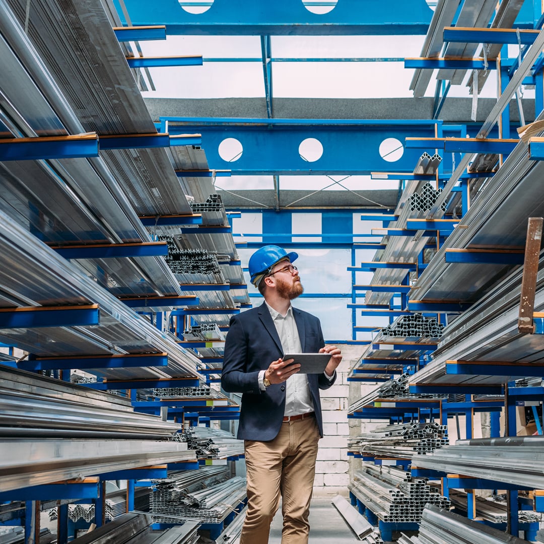 Engineer in a blue hard hat holding a tablet while inspecting rows of steel