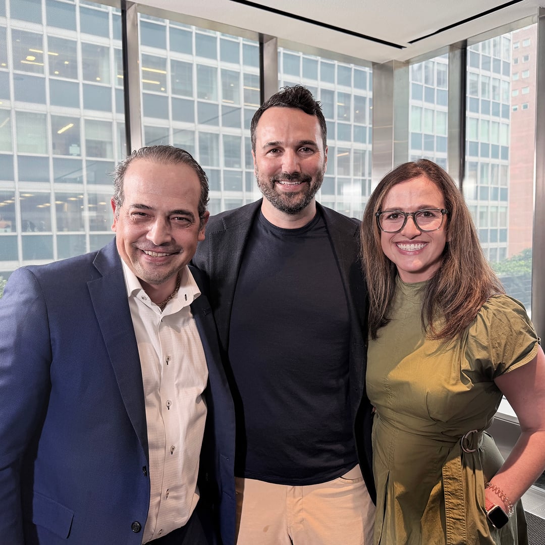 Three smiling business professionals posing together in a modern office with floor-to-ceiling windows
