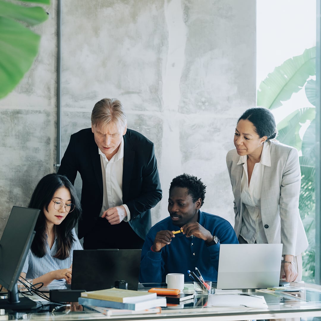 Four diverse colleagues gathered around a desk with laptops in a modern office