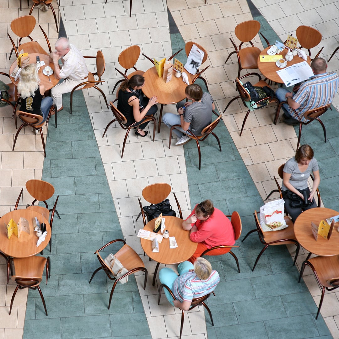 Overhead view of a busy café seating area with small round tables and wooden chairs, where people are seated, eating and talking in a tiled indoor space.