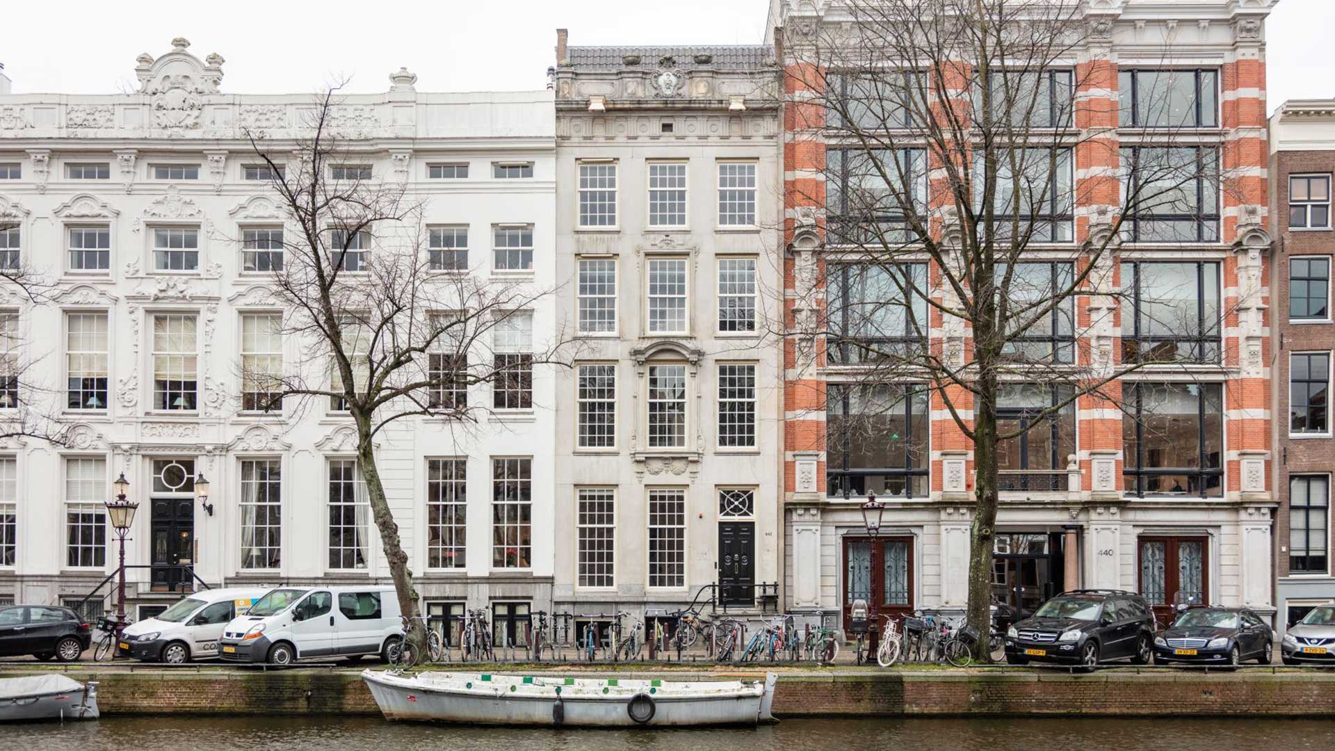 Historische grachtenpanden aan een Amsterdamse gracht met gevels, fietsen langs de kade, geparkeerde autos en een boot op het water.