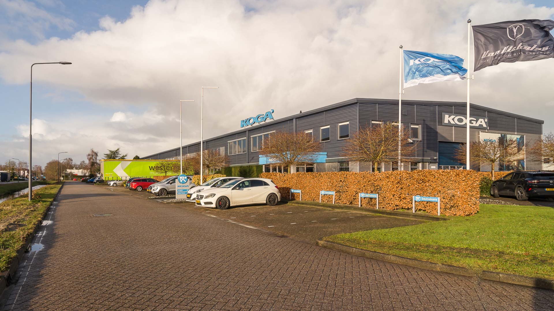 Een modern gebouw met een groot bord waarop 'KOGA' staat. Dit lijkt de locatie van een bedrijf of merk met die naam te zijn. Er zijn verschillende vlaggen met hetzelfde logo die wapperen in de wind. Voor het gebouw is een parkeerplaats met auto’s en fietsen. De lucht is gedeeltelijk bewolkt en het is overdag.