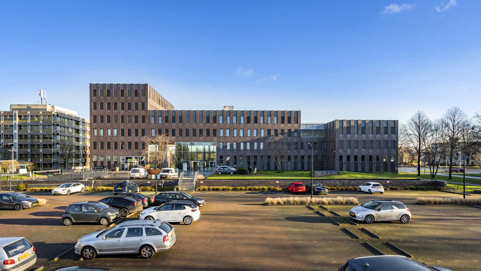 Een modern architecturaal complex met meerdere gebouwen met meerdere verdiepingen die een donkere gevel en rechthoekige ramen hebben. Op de voorgrond is er een parkeerplaats met verschillende geparkeerde auto’s, waaronder een opvallende rode auto. De lucht is helder en blauw, wat suggereert dat het een zonnige dag kan zijn. Deze afbeelding kan interessant of relevant zijn voor discussies over architectuur, stedenbouw, of om het exterieur van dit specifieke gebouwencomplex te laten zien.
