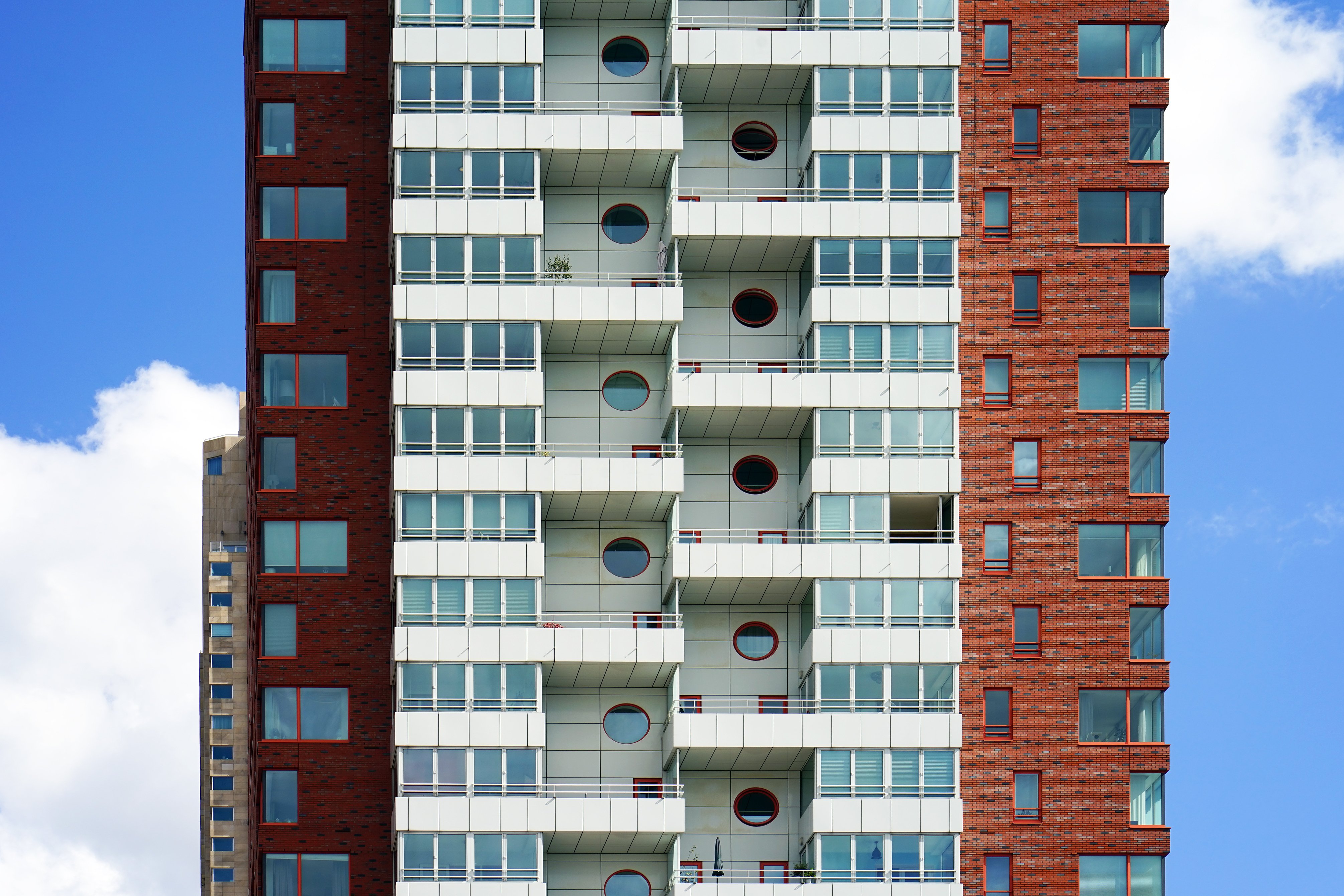 Residential apartment tower with red brick facade, white balconies and large windows. Symmetrical architecture set against a blue sky with clouds.