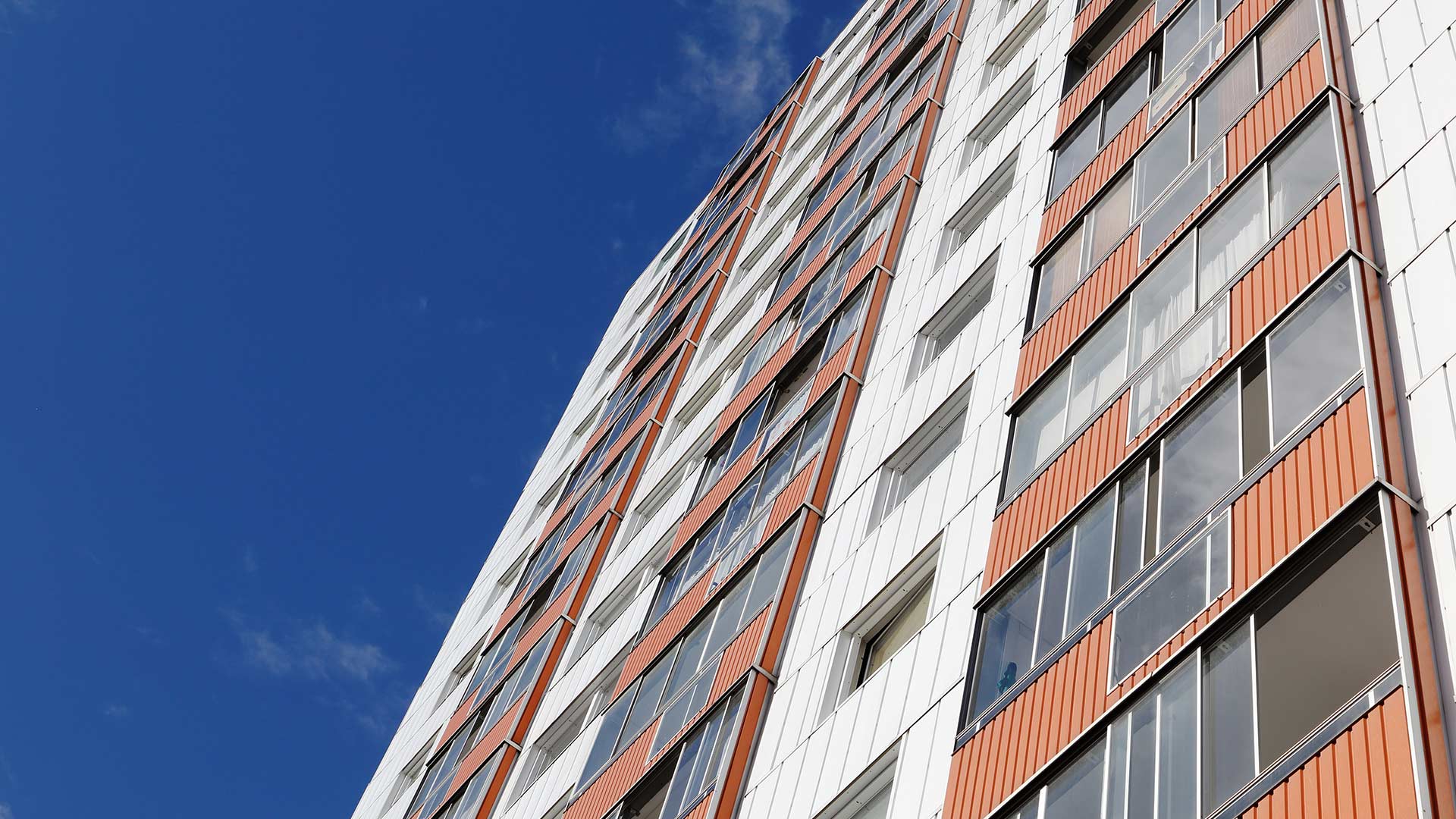 High-rise apartment building with white facade panels and orange brick, viewed from a low angle, showing many windows against a clear blue sky.