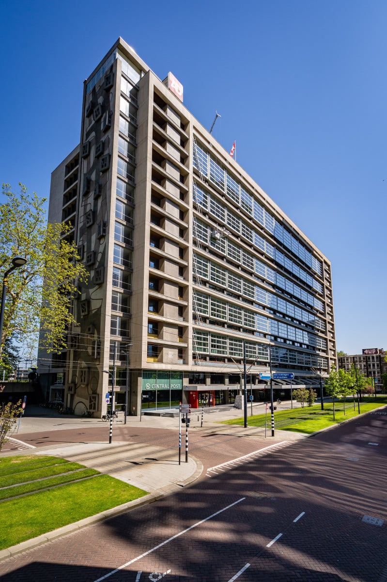 Een gebouw met meerdere verdiepingen, glazen gevel, balkons en winkels op straatniveau, omringd door bomen in een stedelijke omgeving.
