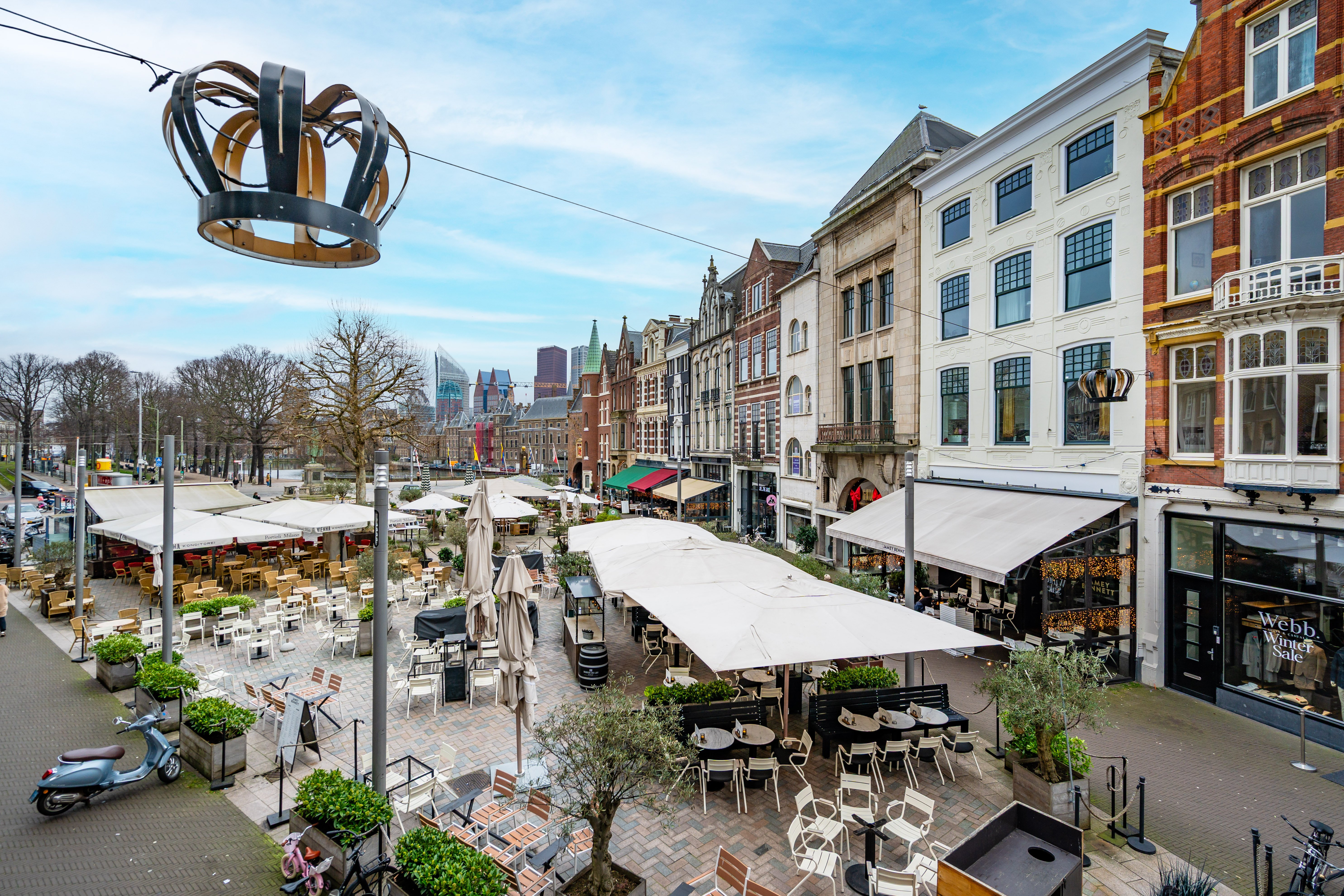 Lively square with cafe terraces and umbrellas, surrounded by historic facades and shops, hanging crown decoration above street and urban skyline in background