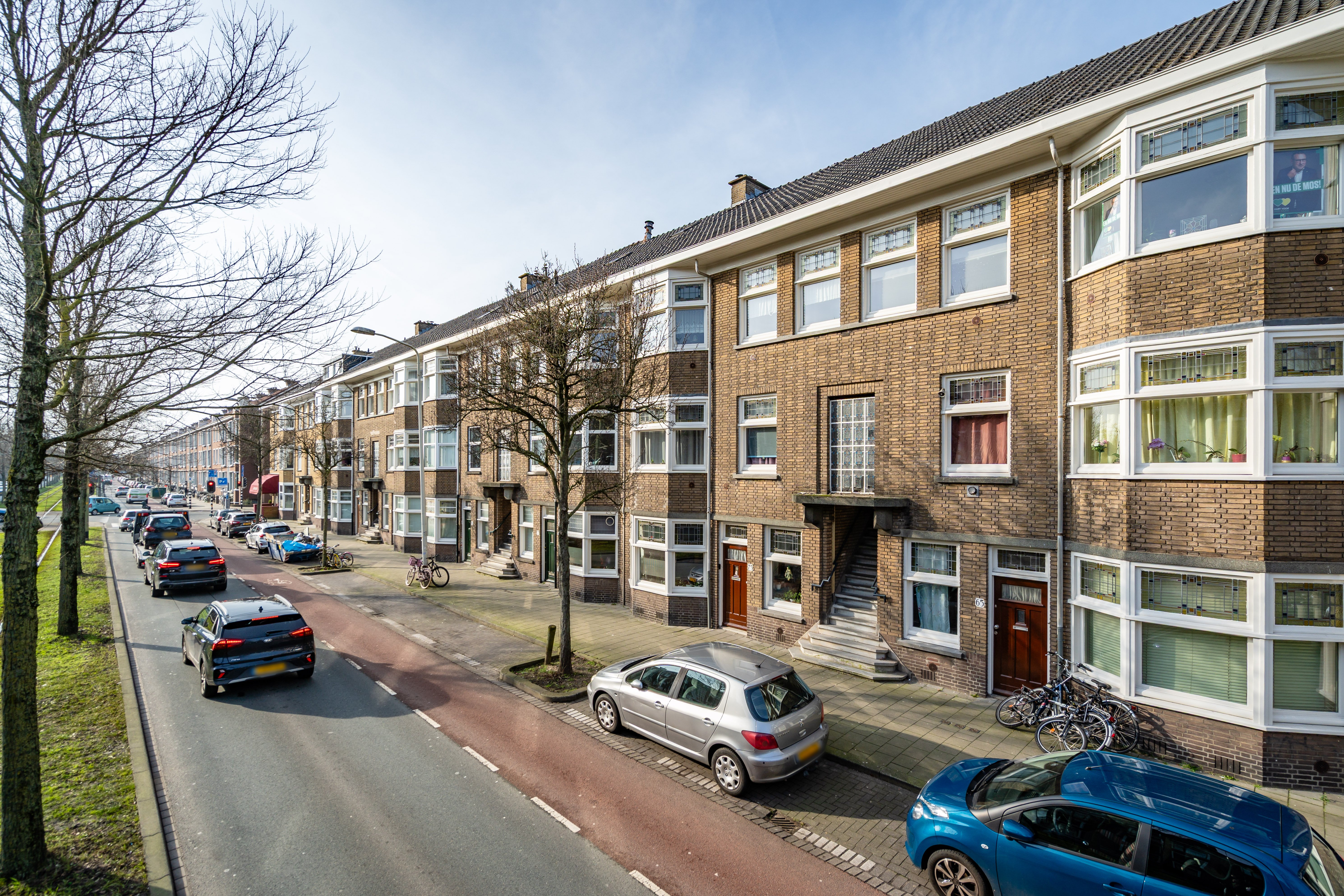 Street view with row of brick walk up apartments, parked cars along roadway and bike lane, leafless trees and wide sidewalk in quiet residential area under clear sky