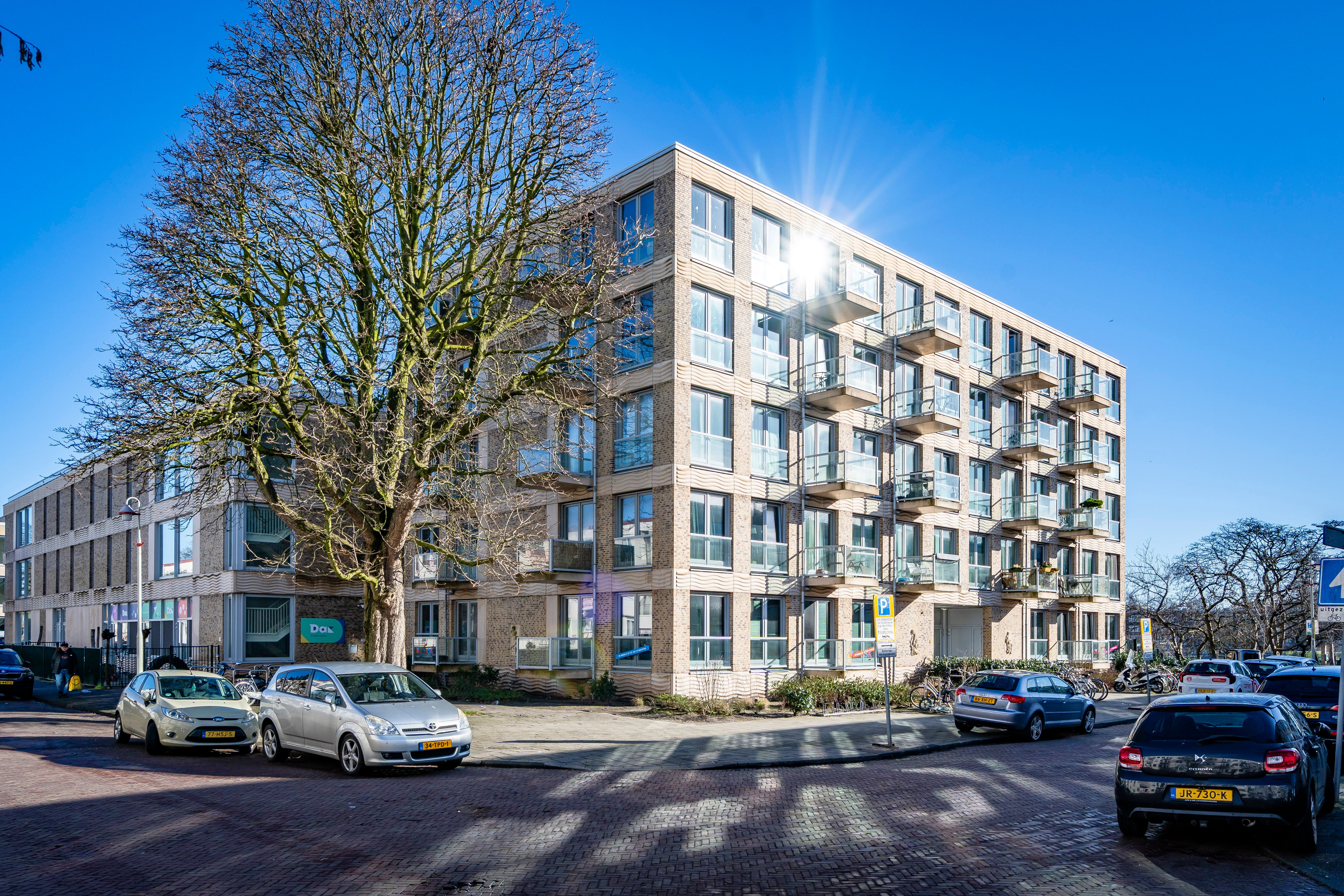 Corner location with brick apartment building of five floors, balconies and large windows, tree line and parked cars along street under clear blue sky with sun reflection