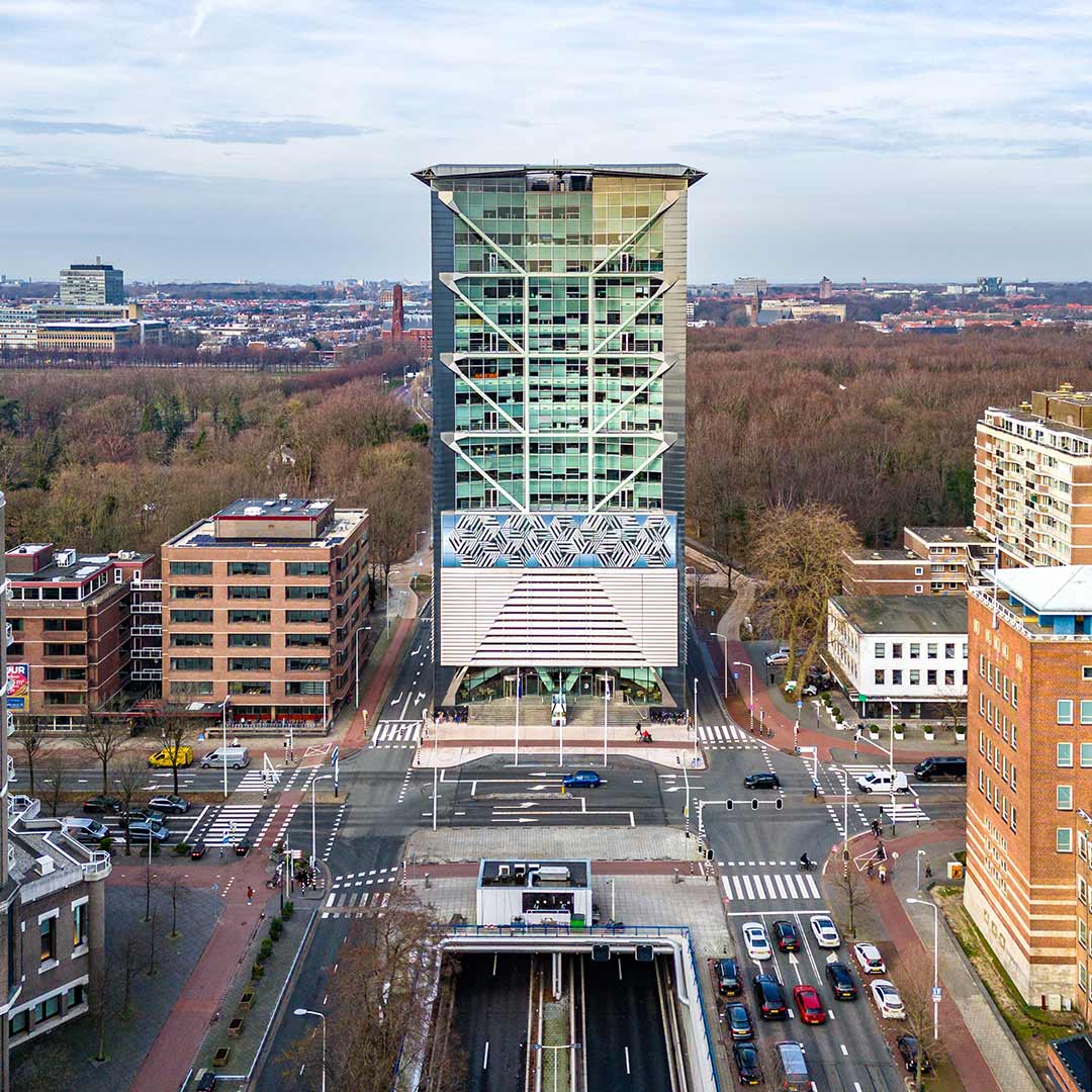 Luchtfoto van de Malietoren in Den Haag aan Bezuidenhoutseweg, omringd door kantoorgebouwen, woningen, kruispunt en stedelijk groen met bosgebied op de achtergrond.