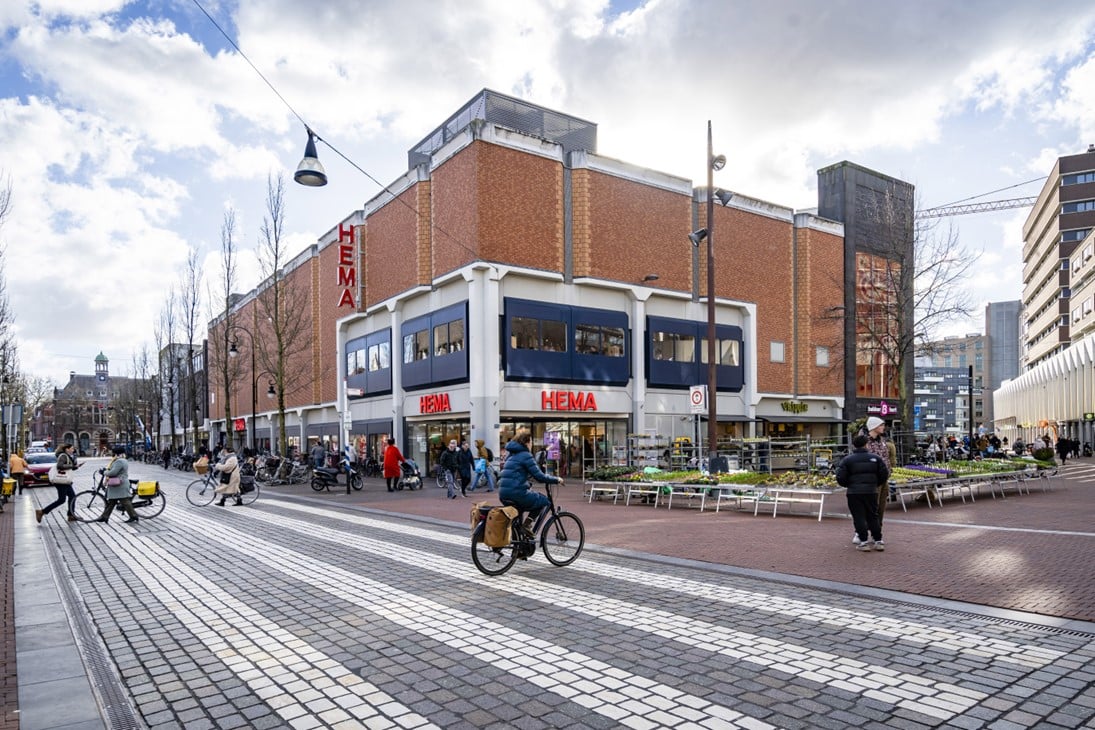 Straatbeeld met HEMA warenhuis op een hoekpand, fietsers en voetgangers bij een kruising, winkelgebied in een stedelijke omgeving met terrassen.